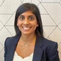 Professional woman with black hair, wearing a navy blazer and white top, smiling, with a geometric wall background.