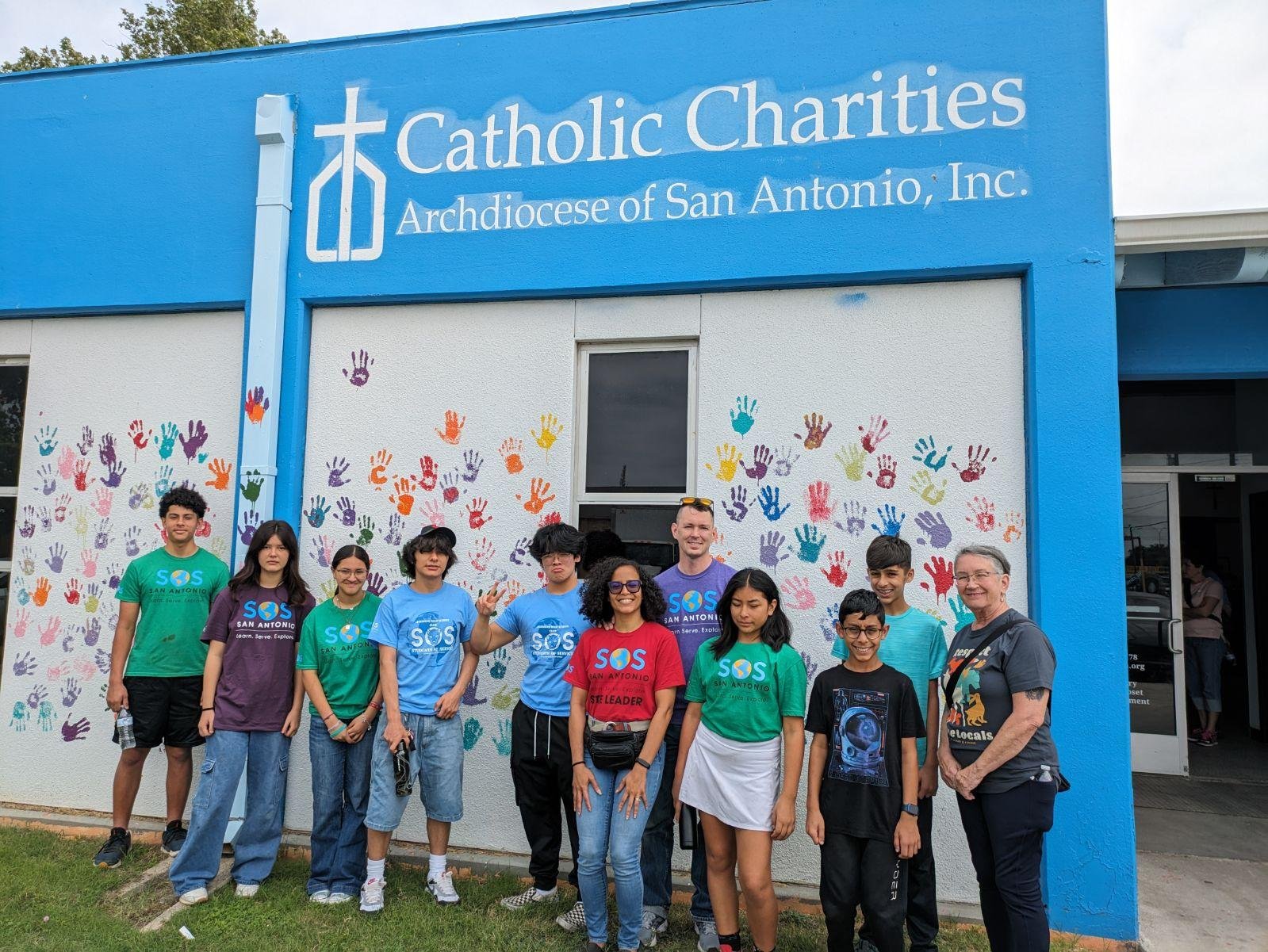 Group of people standing in front of a building labeled 'Catholic Charities, Archdiocese of San Antonio, Inc.' with colorful handprints on the wall behind them.
