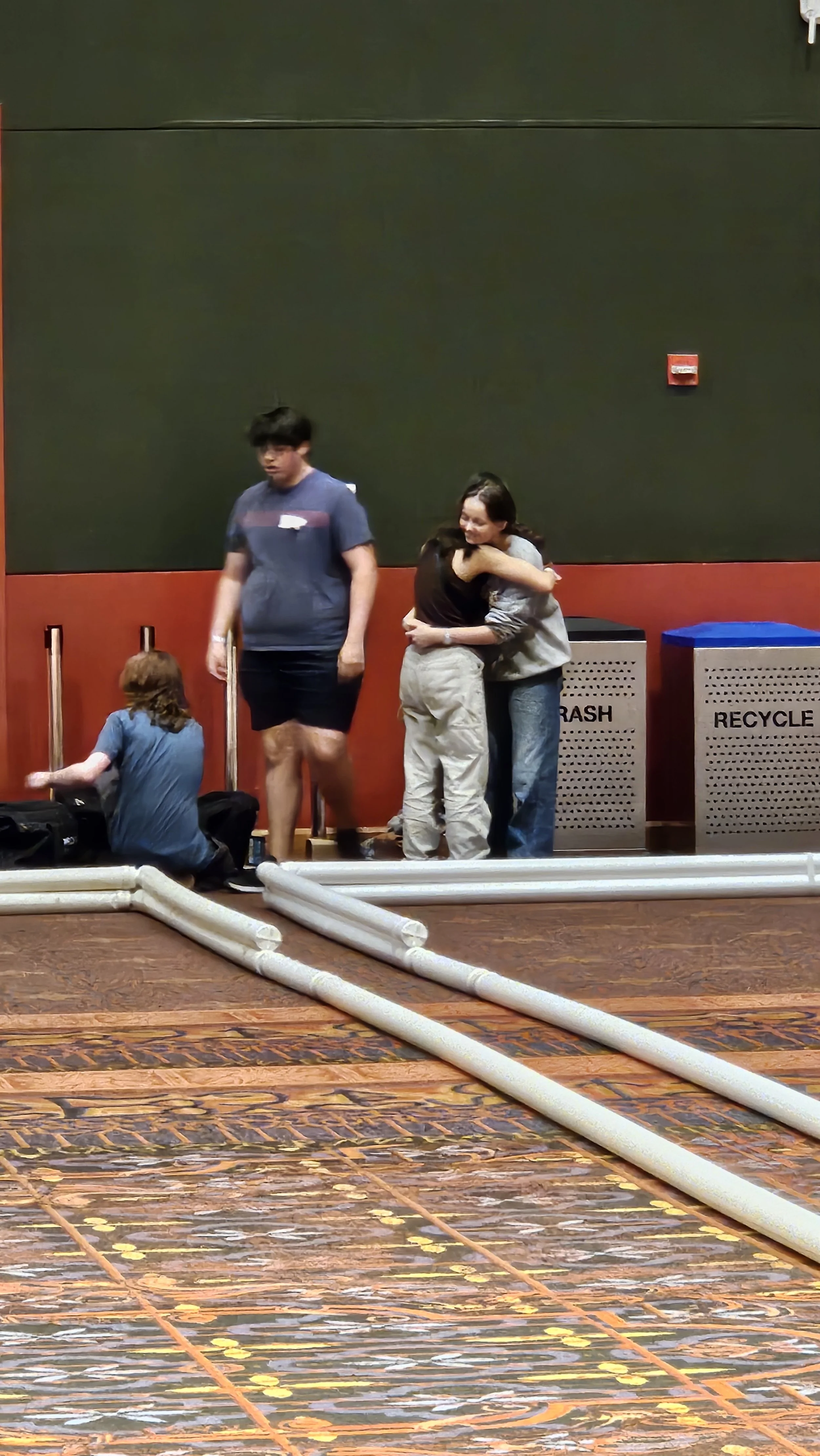 Two women hugging each other while a man stands nearby in an indoor setting near recycling bins and pipes.