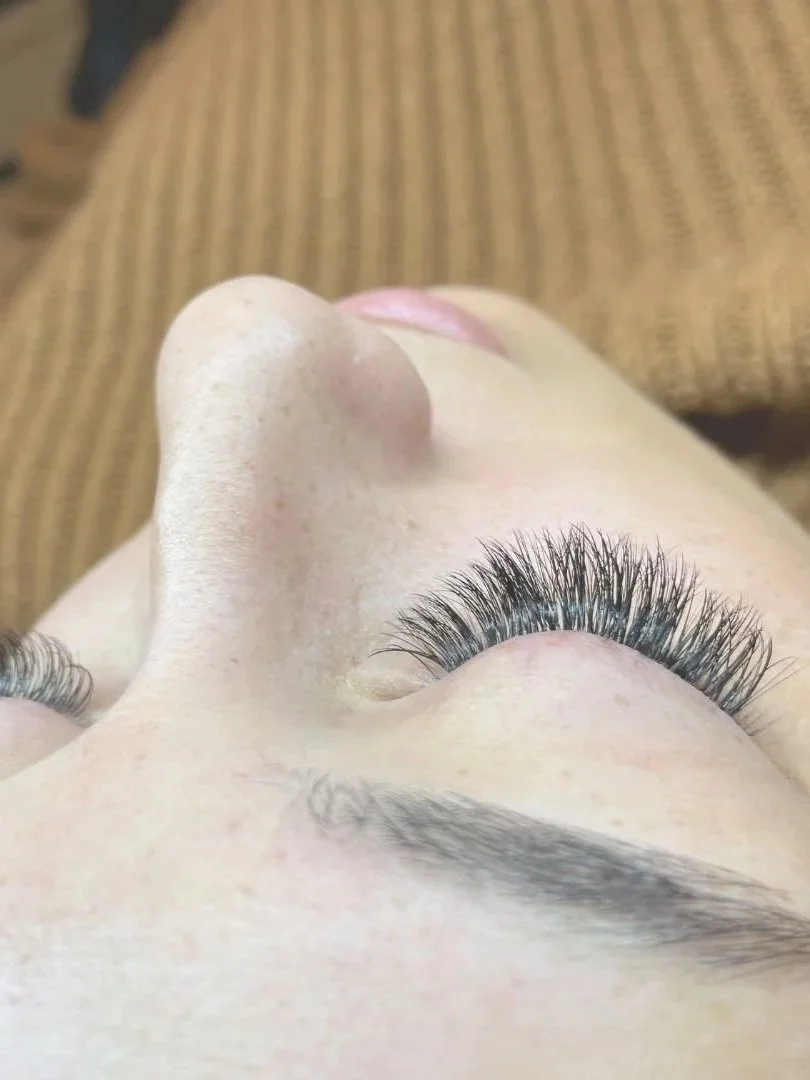 Close-up of a person's face from above, showing eyelash extensions on closed eyes and a piece of furniture in the background.