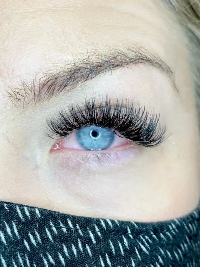 Close-up of a blue human eye with long, dark, voluminous eyelashes and partial view of an eyebrow, with the person wearing a black face covering.
