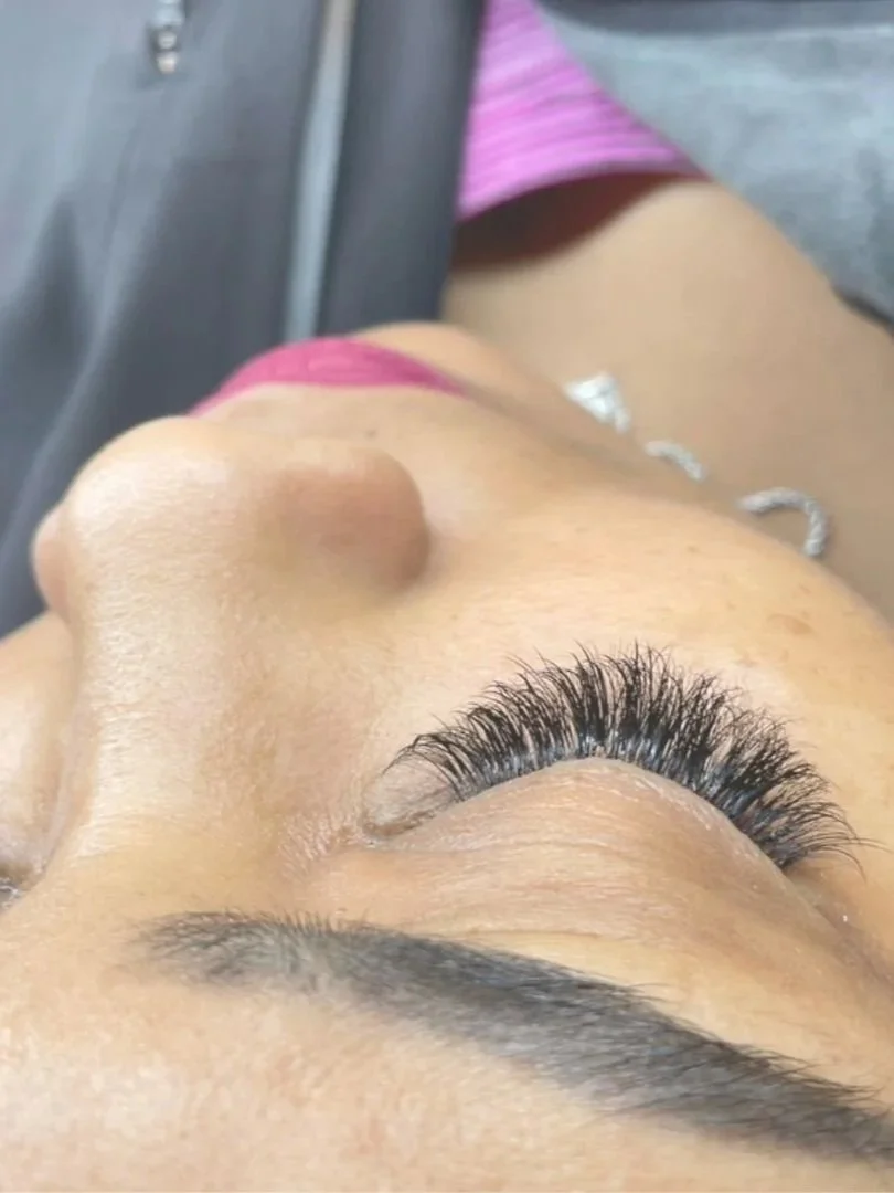 Close-up of a woman's face showing her face with long, curled eyelashes, and her eyes closed. She has dark eyebrows, smooth skin, pink lipstick, and is wearing a silver necklace and pink clothing.