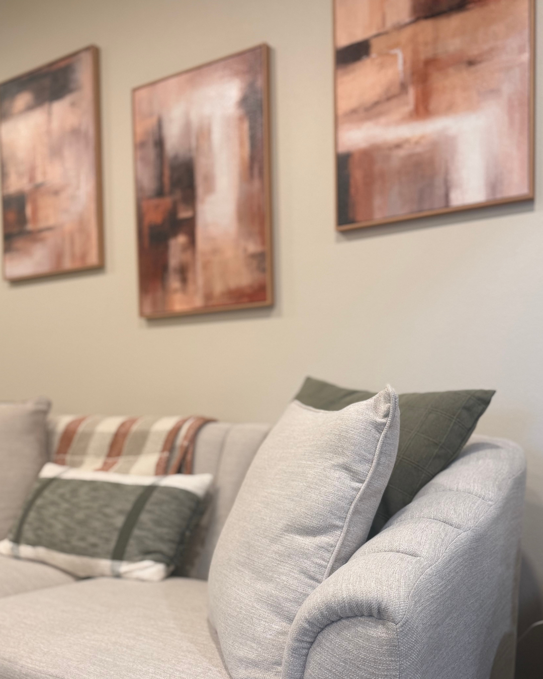 A relaxation room with beige sofa, patterned cushions, wall-mounted shelves, abstract artwork, potted plants, and a corner of a black cabinet or fireplace.