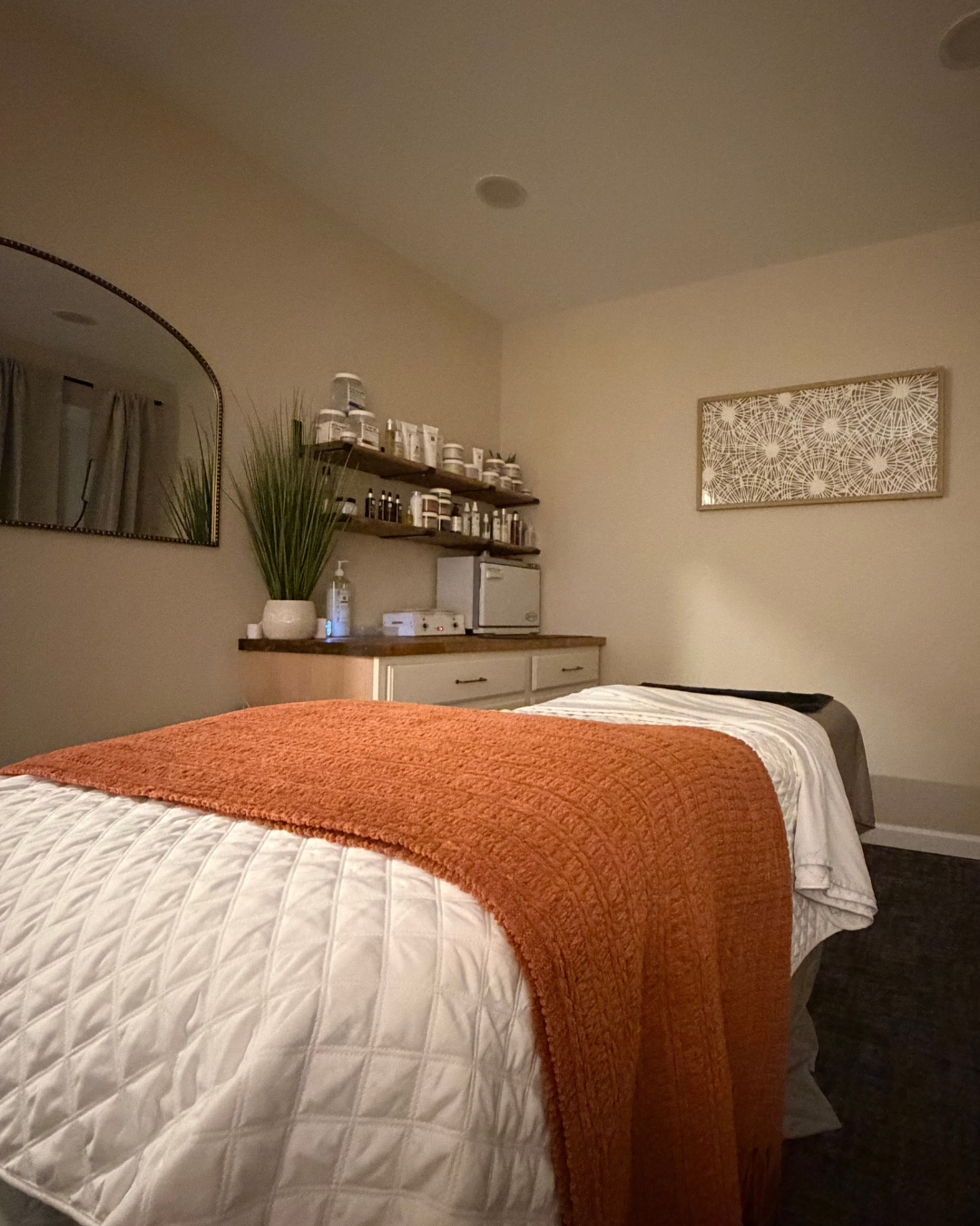 A facial treatment room with a bed covered in a gray and white quilt, a wall with two framed pictures, a white cabinet with shelves holding toiletries, and a small trash can.