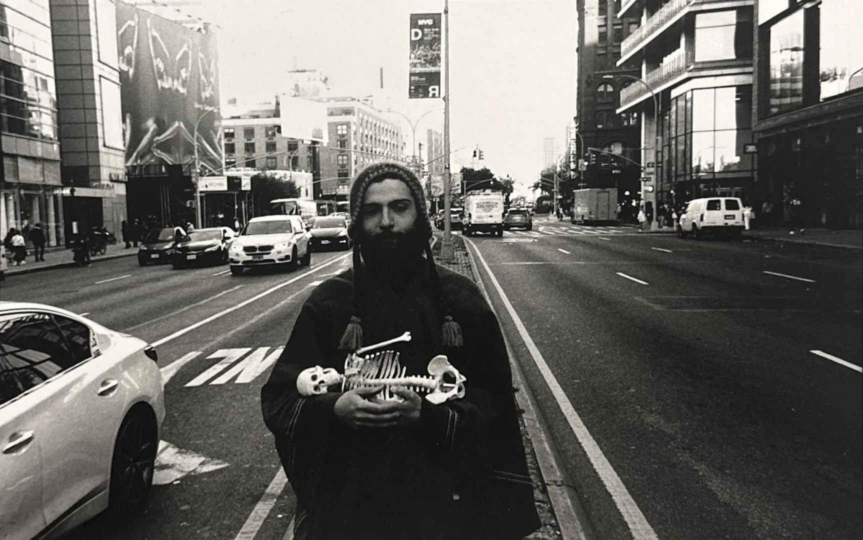 A person standing on a city street holding a small skeleton in their hands. The street is busy with cars and buildings on both sides.