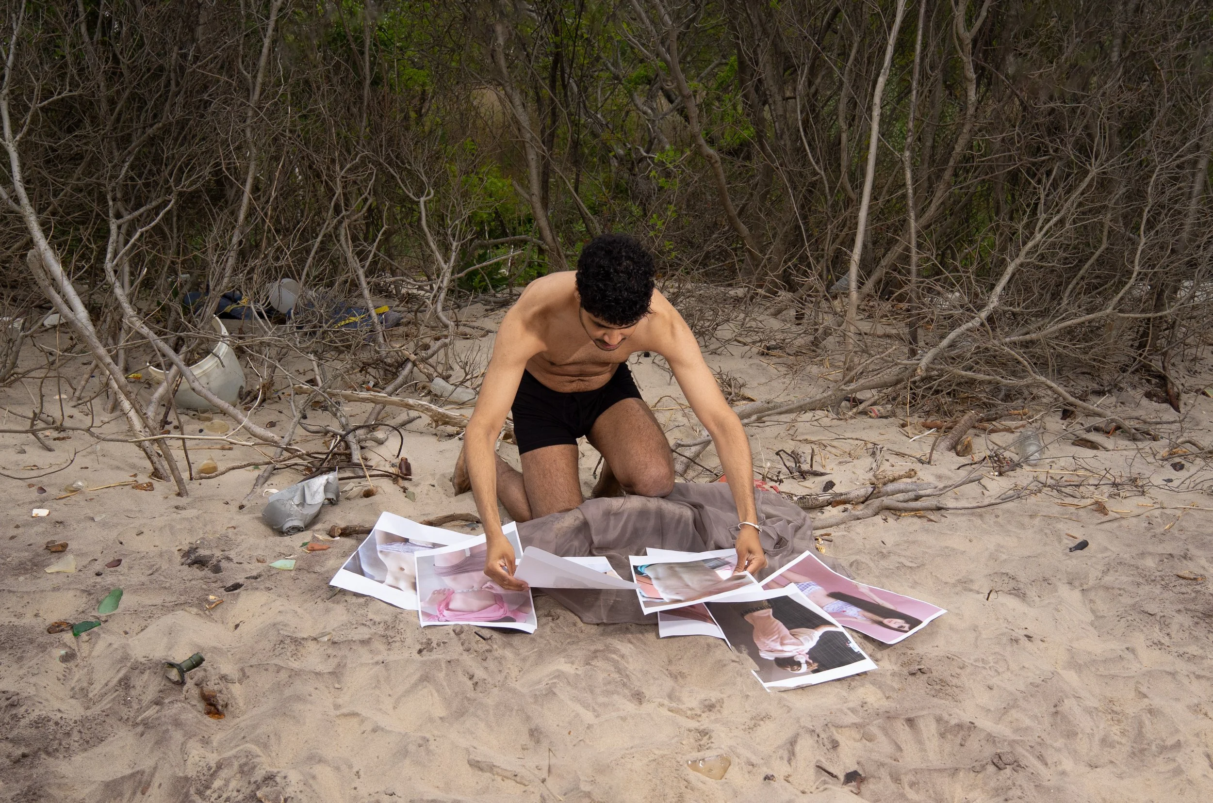 Man crouching on sandy beach, surrounded by scattered photographs and debris, with leafless bushes in the background.
