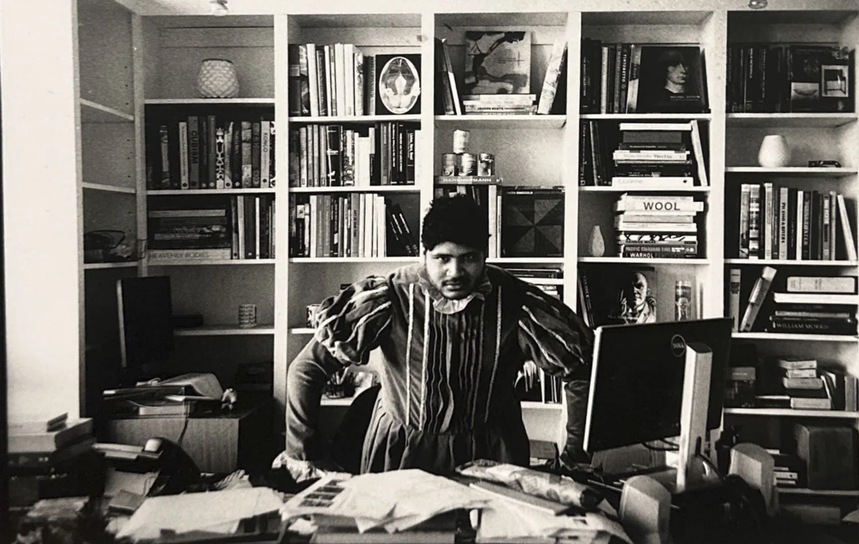 Black and white photo of a man wearing a striped shirt, sitting at a cluttered desk in front of a bookshelf filled with books and decorative objects.