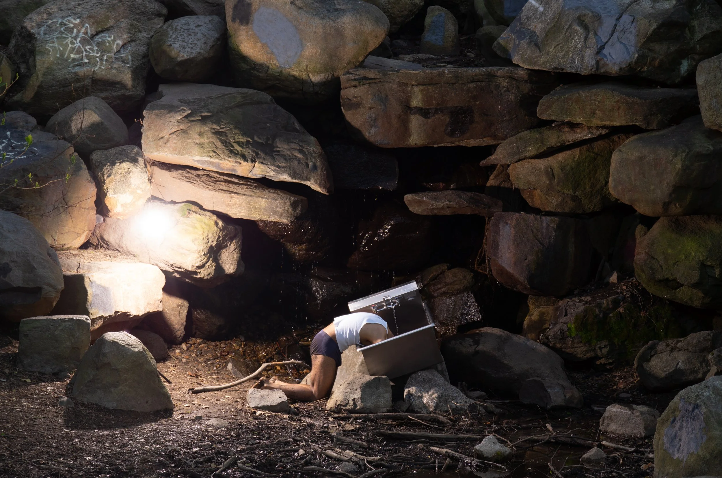 Person wearing white shirt and dark underwear bowing down with head and upper body inside a metal container, surrounded by large rocks in a rocky outdoor area.