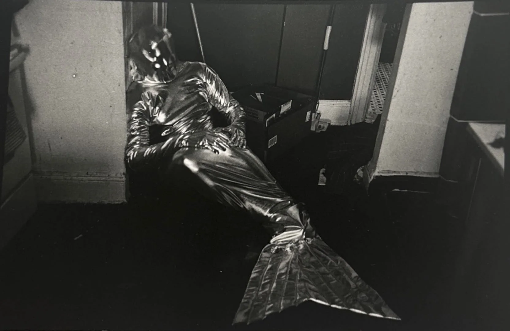 A mermaid with a shiny, metallic tail and top, sitting on the floor in a room with cabinets and boxes, resting or sleeping with eyes closed.