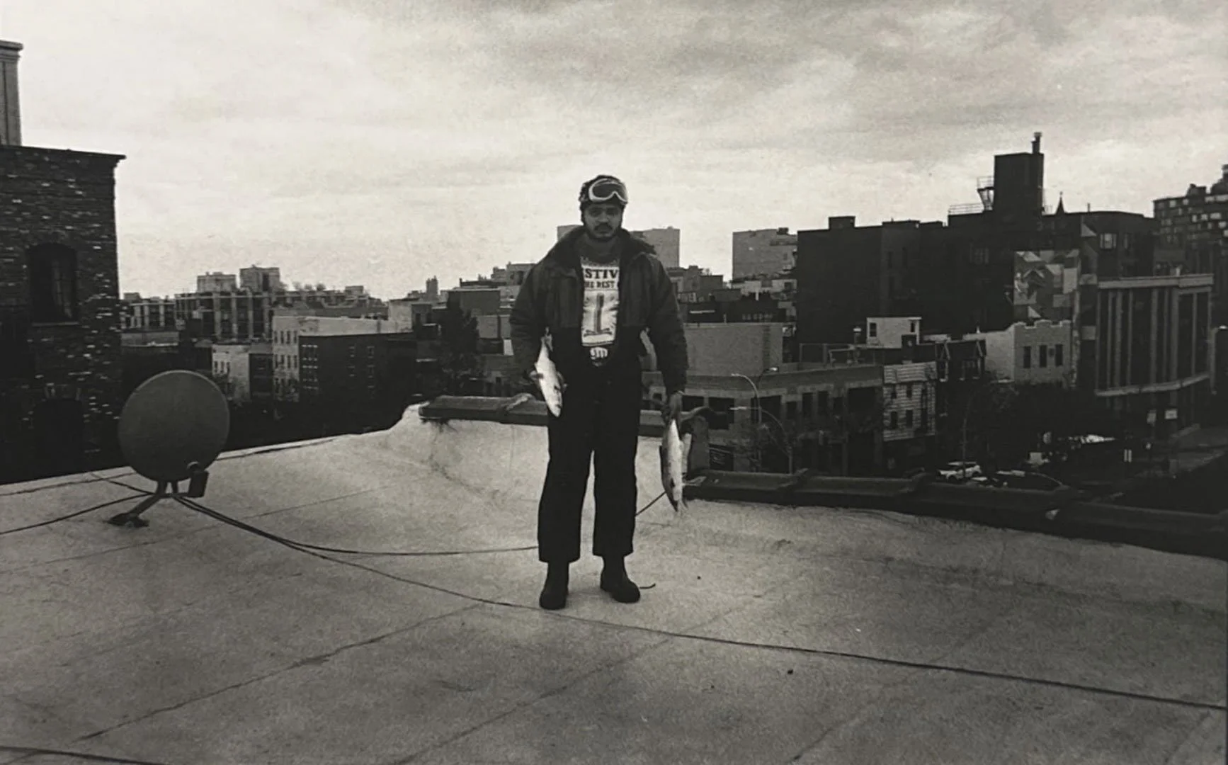 A man standing on a rooftop holding two fish, wearing sunglasses, a jacket, and dark pants, with a cityscape in the background.