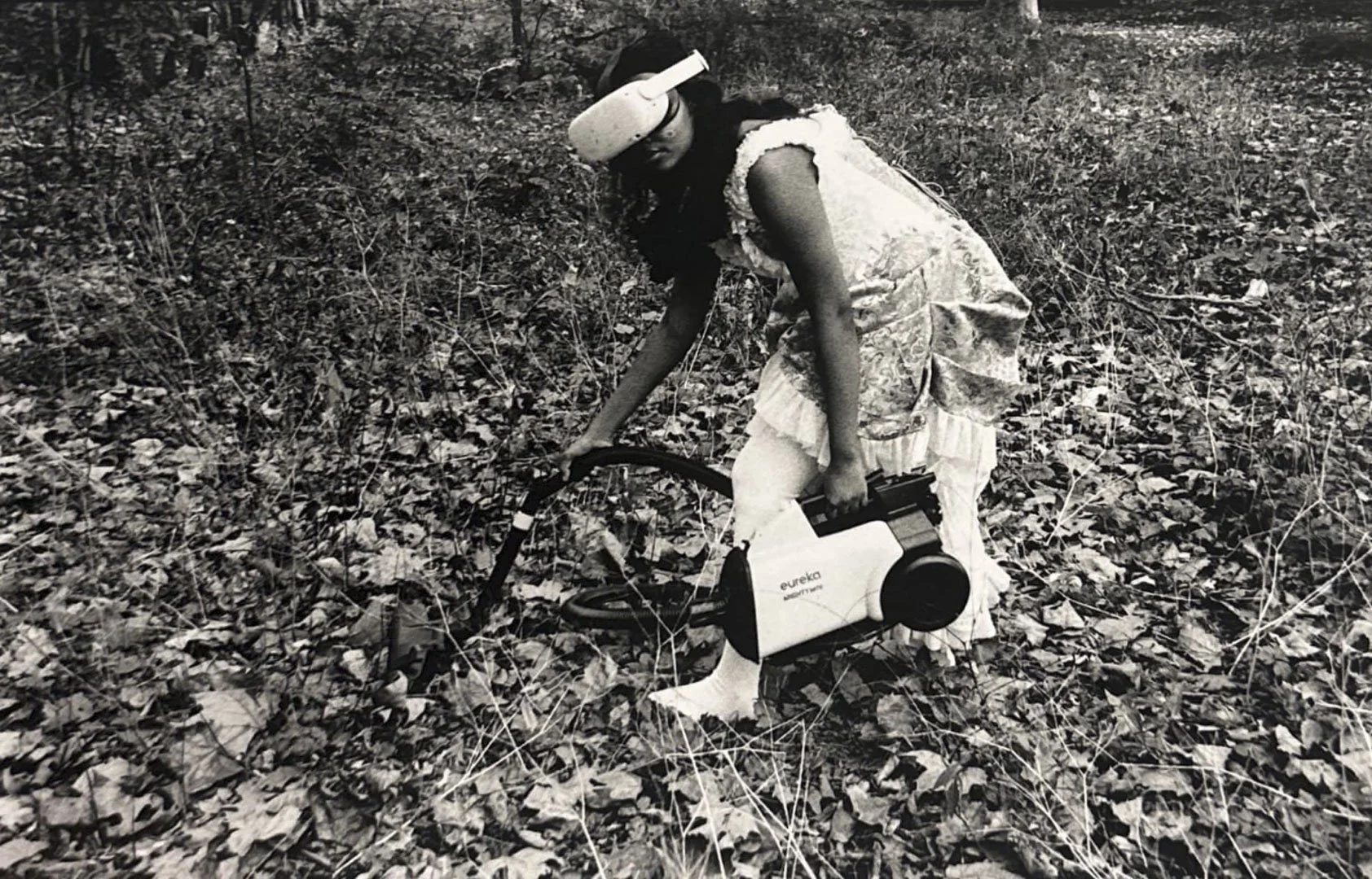A young girl in a dress and shirt playing outside in the woods, holding a small vacuum cleaner.