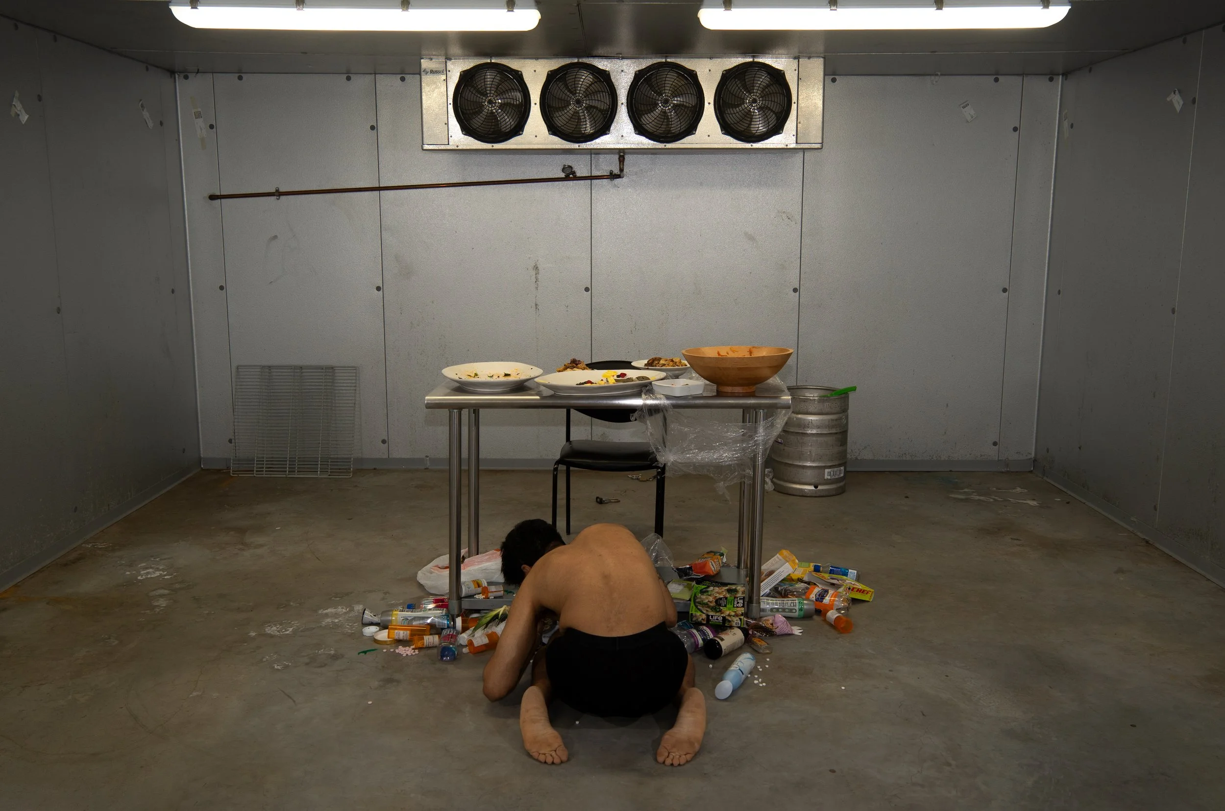 A person crouched on the floor amid trash with spilled pills, bottles, and wrappers in a stark, industrial-looking room. The room contains a table with dirty dishes and an empty chair.