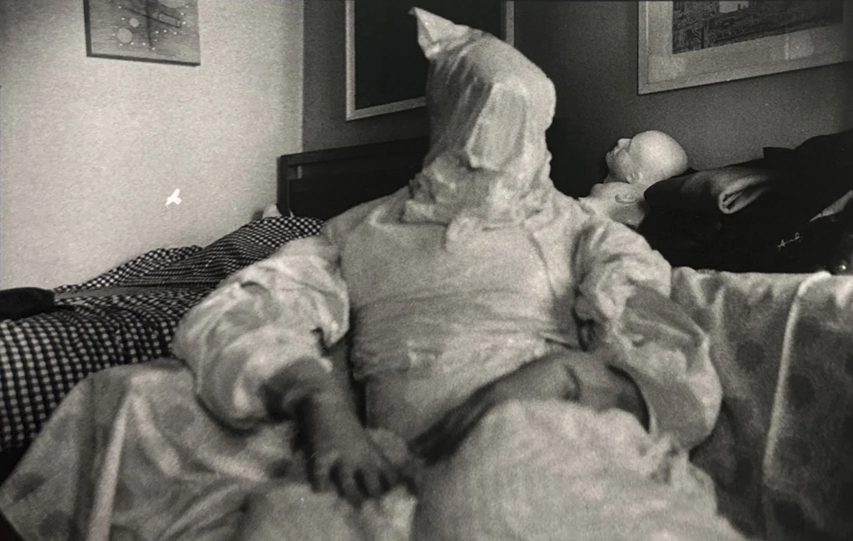 A person sitting on a bed, wearing a full body covering, in a bedroom with paintings on the wall.