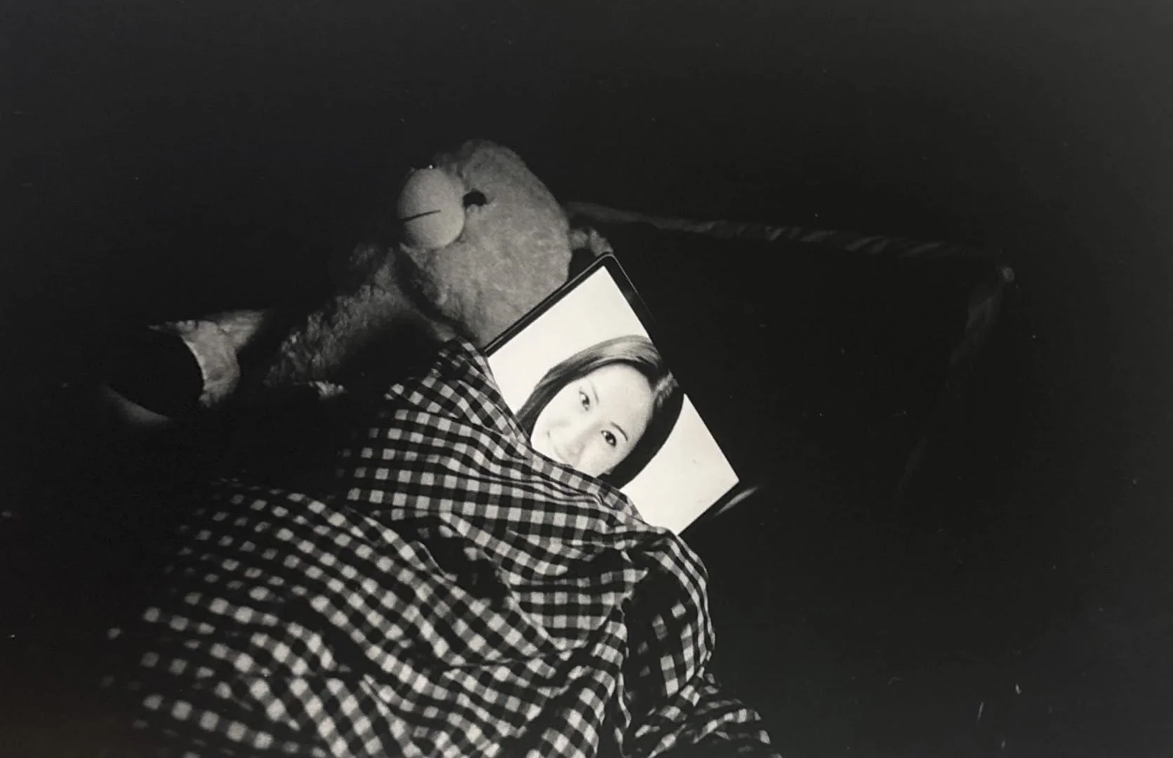 A person lying on a checkered blanket with a teddy bear and a tablet displaying a monochrome photo of a woman with dark hair.