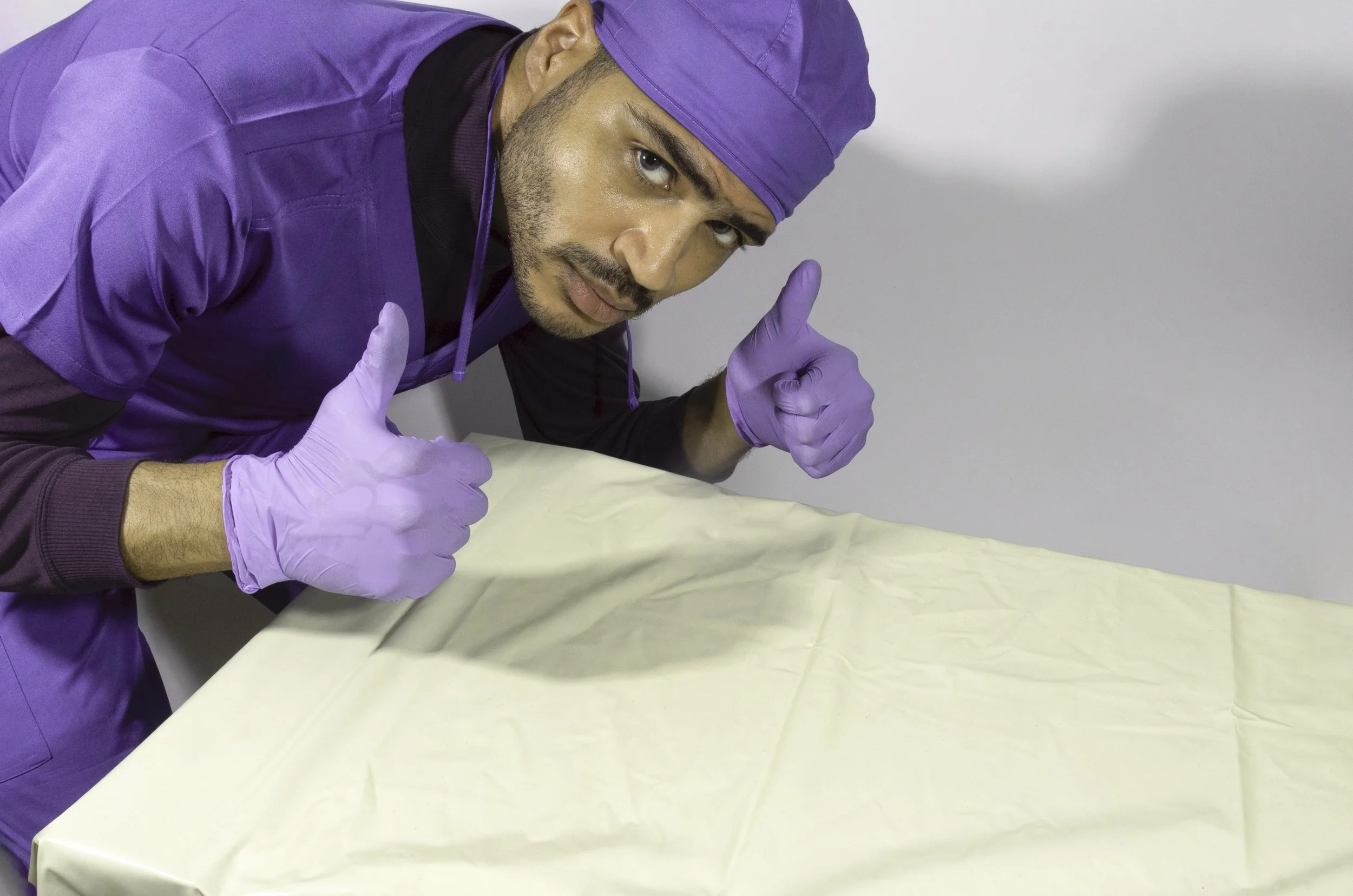 A man in purple medical scrubs and purple gloves giving a thumbs-up gesture while leaning over a table with a light-colored cover.