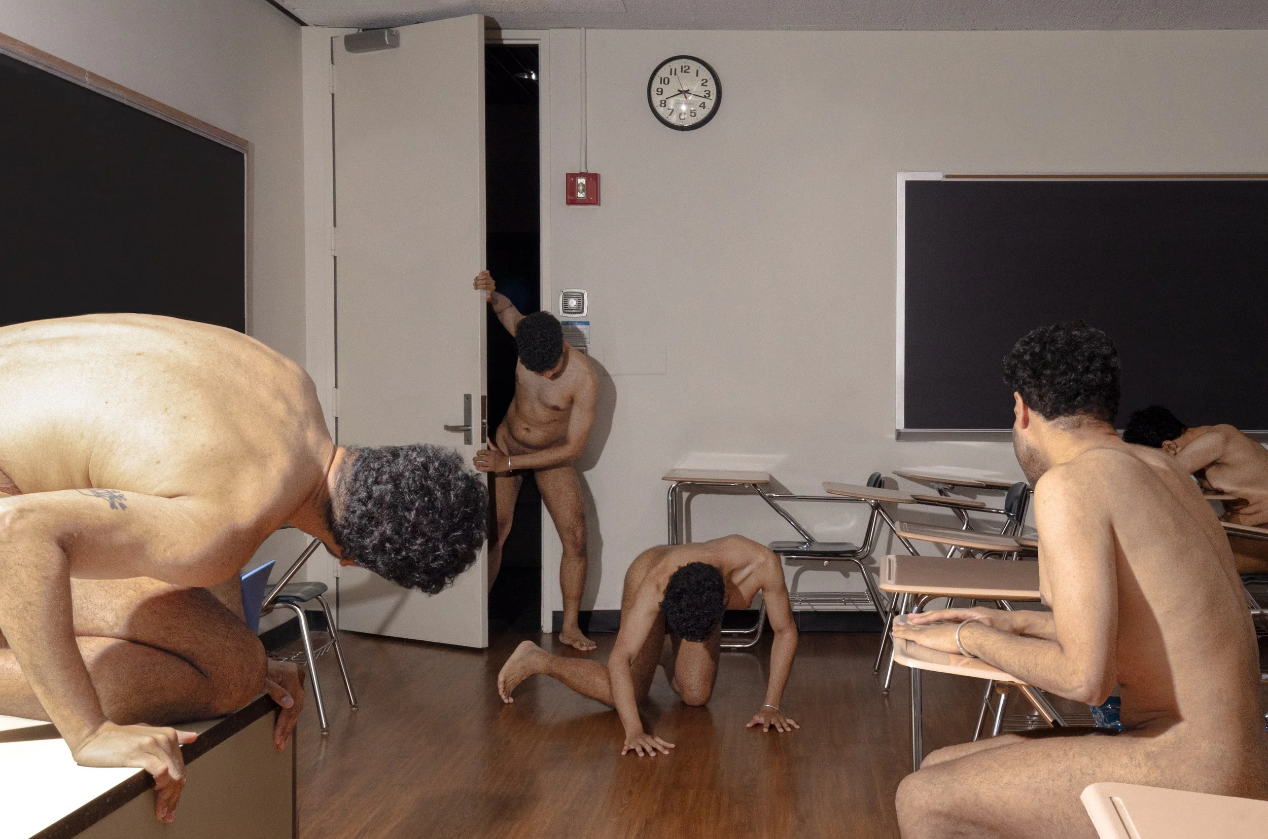 A classroom with four naked men in various positions and one man partially visible entering through a door. The classroom has desks, chairs, a blackboard, and a wall clock showing 2:15.