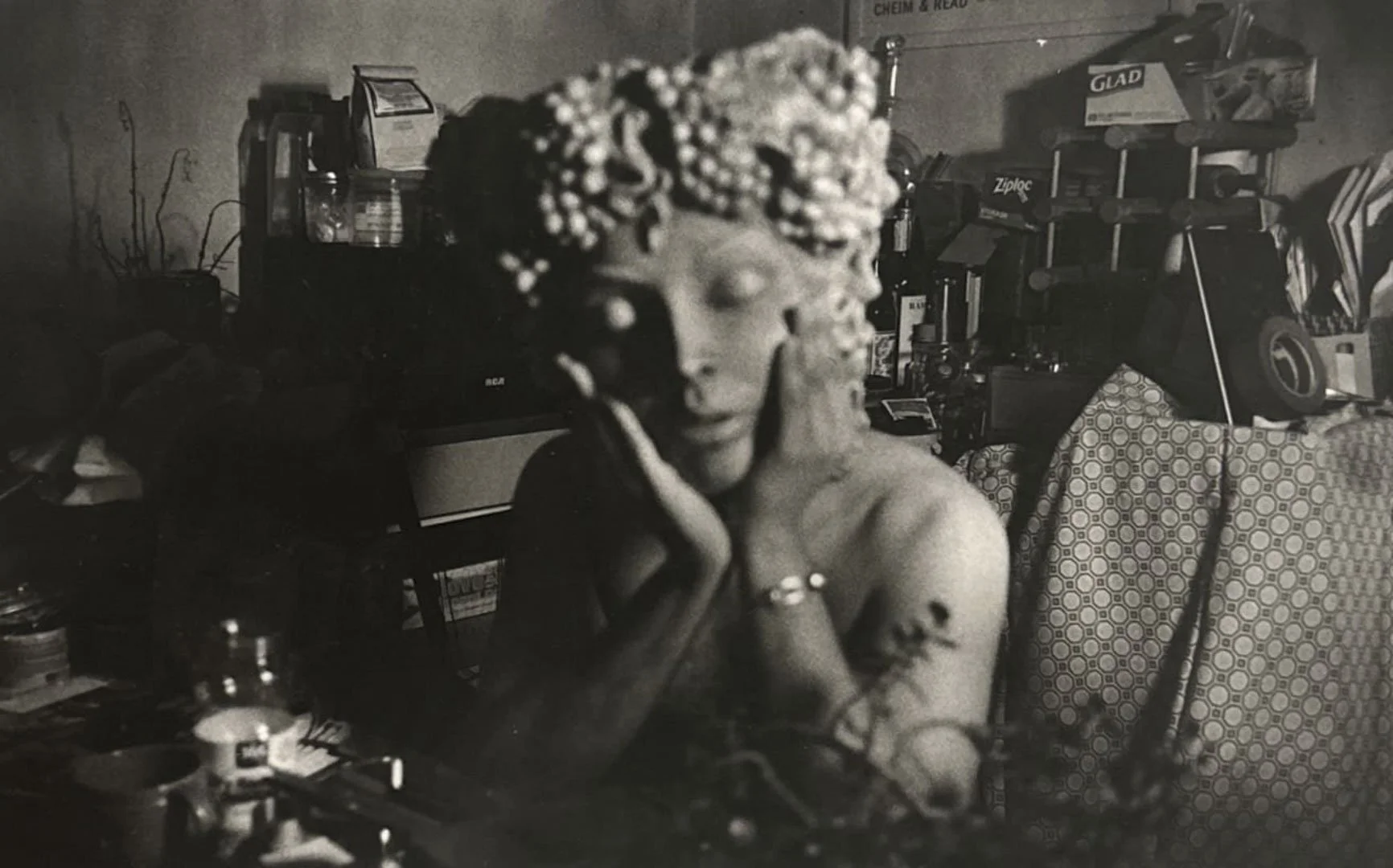 A woman with curly hair adorned with beads, resting her head on her hand, seated in a cluttered room with various objects and shelves in the background.