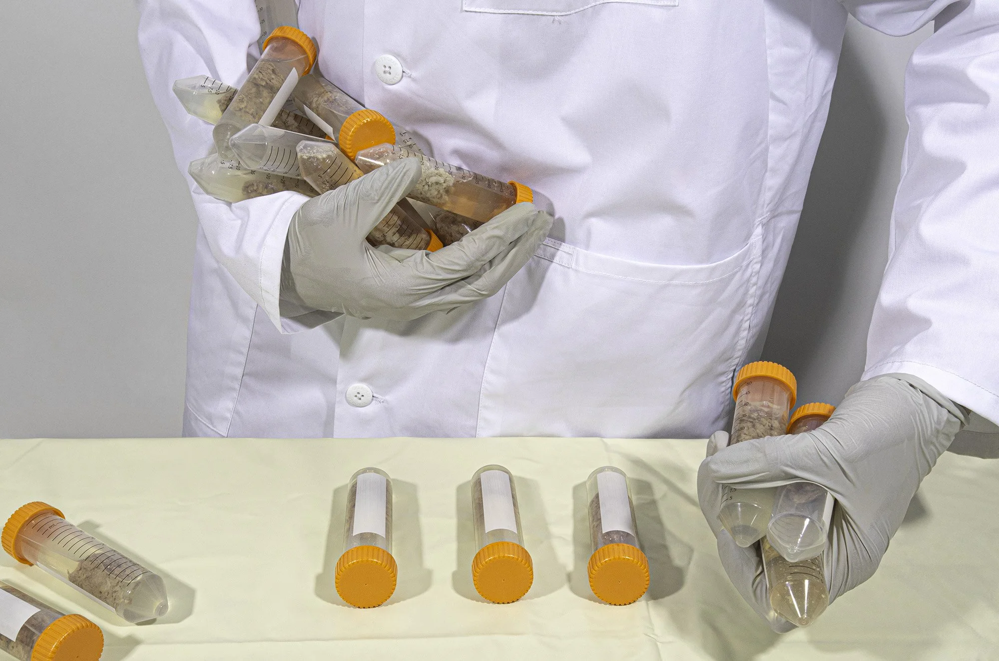 Person wearing white gloves and a white lab coat holding several specimen collection tubes with orange caps, with more tubes laid on a table below.