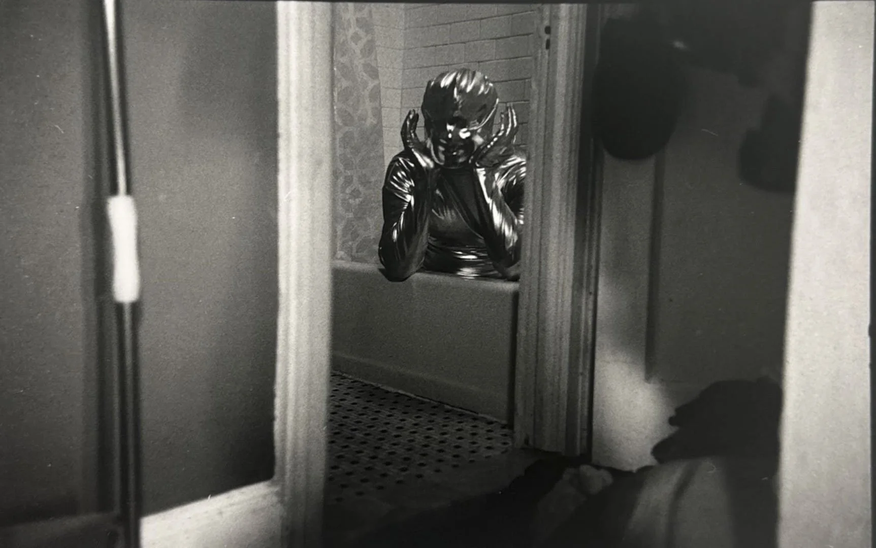 A person wearing a shiny, metallic mask and black leather outfit is looking into a mirror, resting their face in their hands.