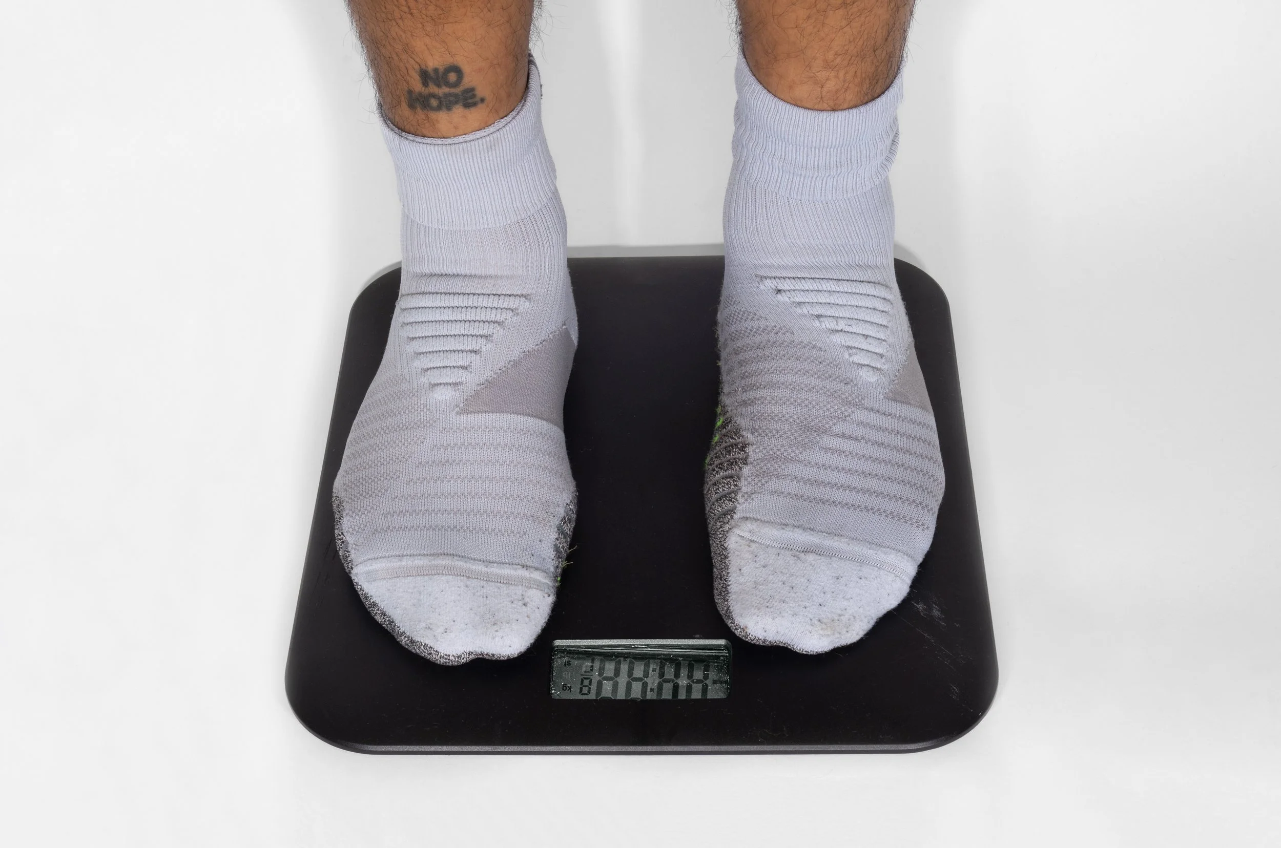 A pair of feet in white athletic socks on a digital scale, displaying an obscured weight. The person has a tattoo on the right calf that reads 'NO HOPE'.