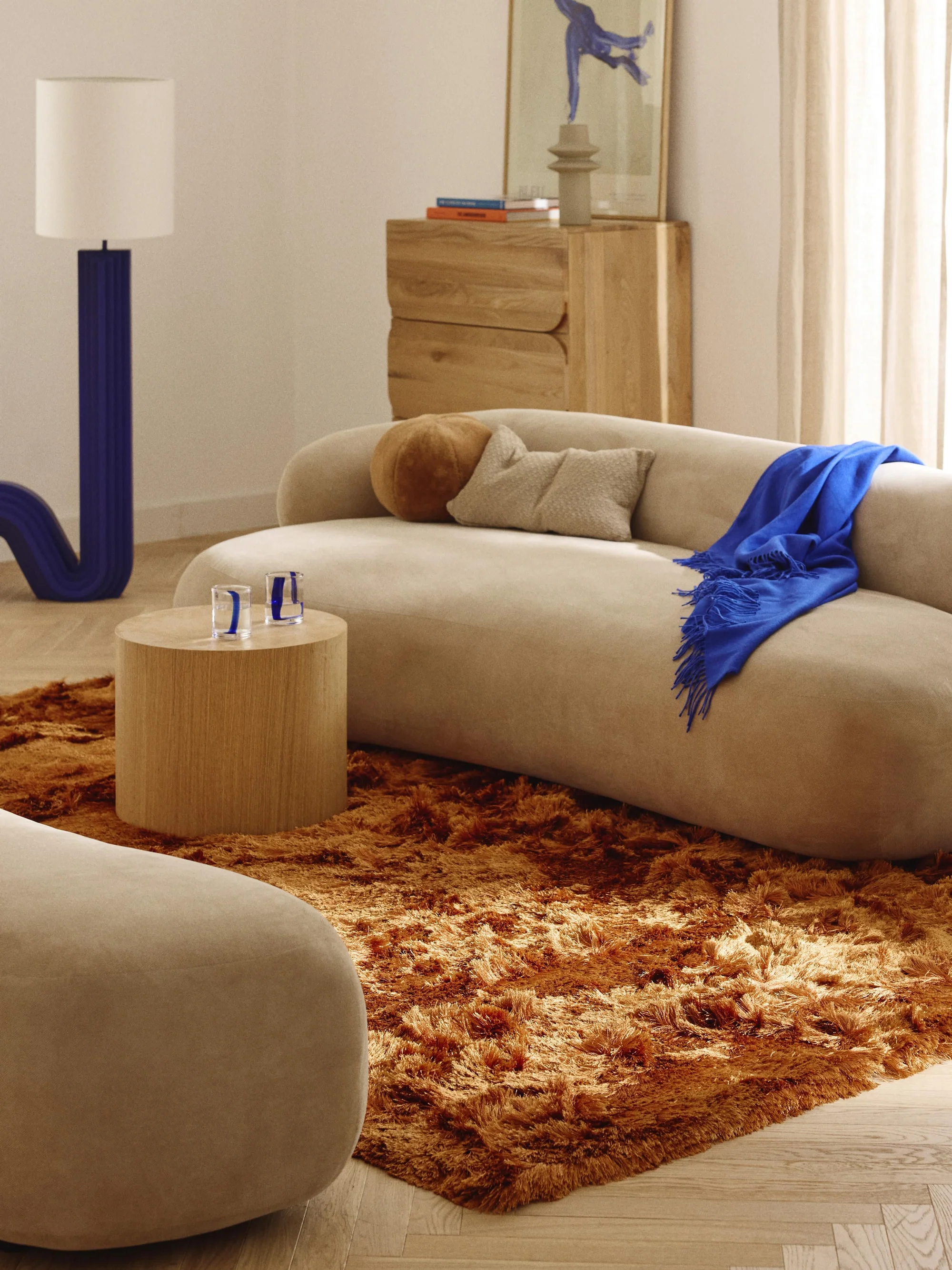 Salon moderne avec canapé beige, tapis en peluche marron, table ronde en bois clair, lampe avec base bleue, décoration artistique sur le mur, rideaux clairs, et coussins et jeté bleu sur le canapé.