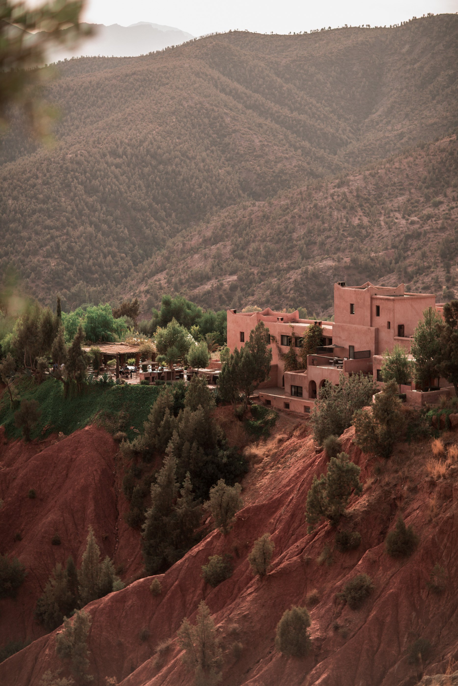 Maisons en adobe rose sur une colline rocheuse dans un paysage montagneux aride, avec des arbres et une zone verte en bas.