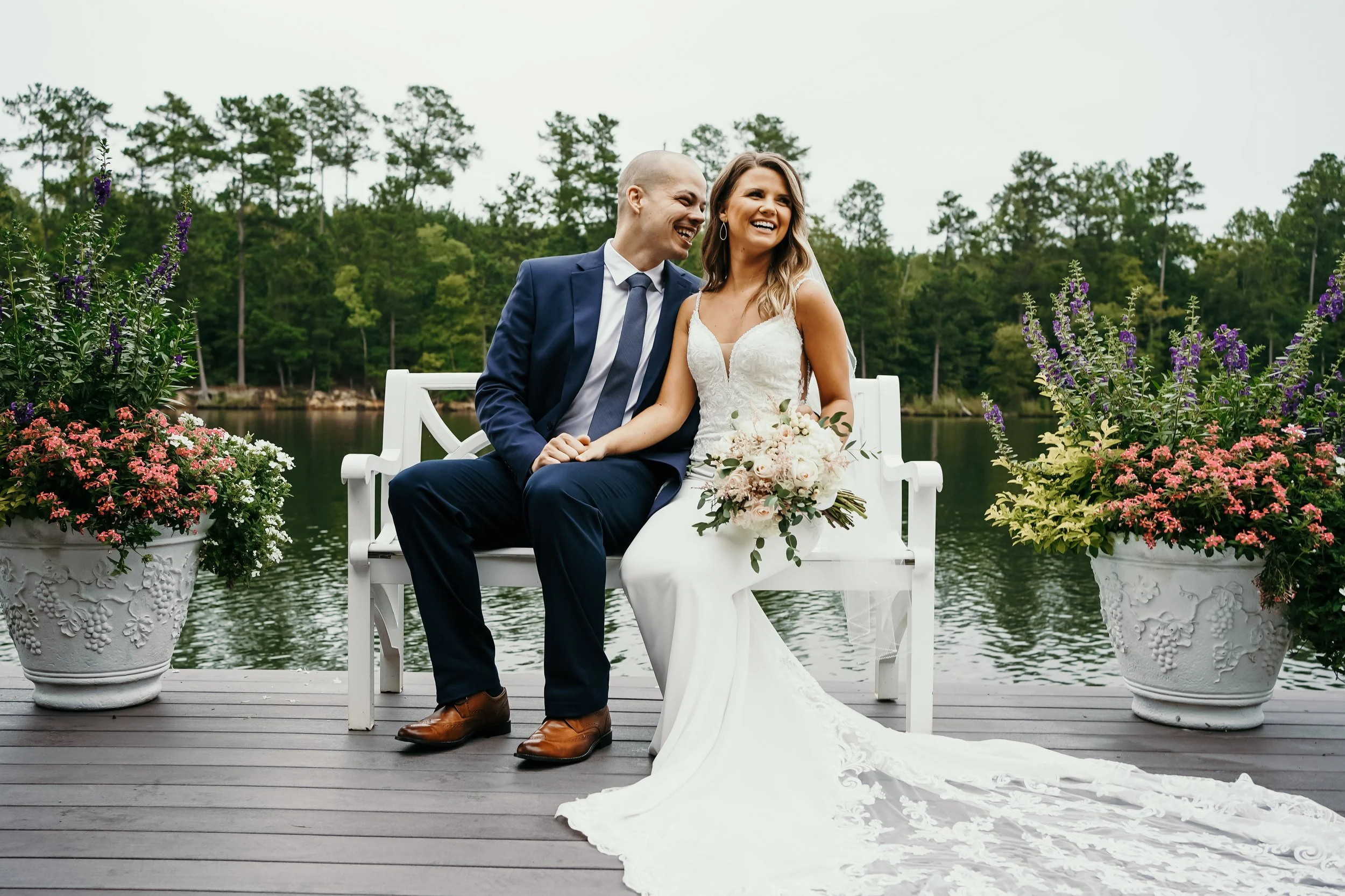 A bride and groom sitting on a white bench by a lake, smiling and laughing, with large flower pots containing pink and purple flowers on either side, and a wooded area in the background.