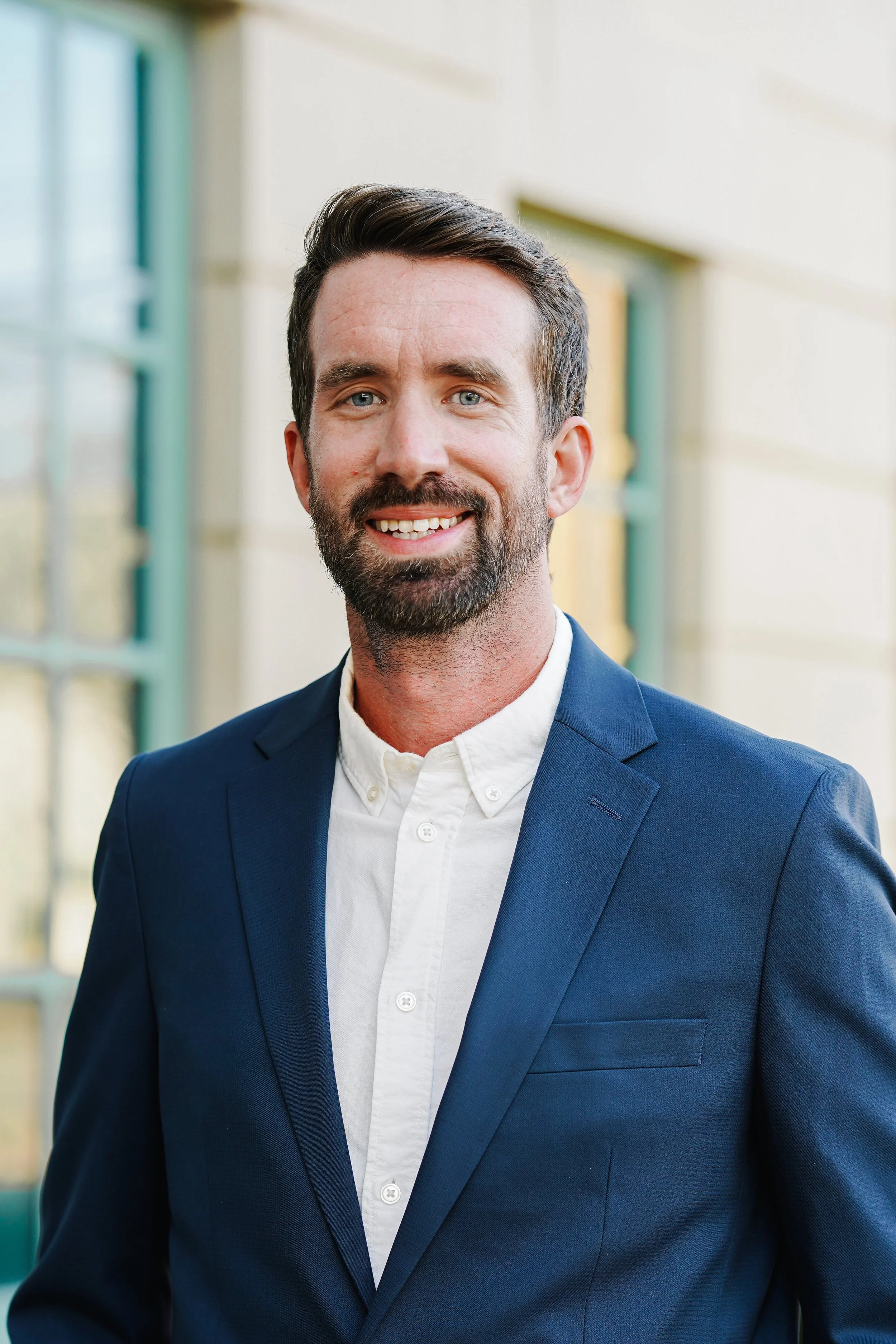 A smiling man with dark hair and a beard wearing a blue blazer and white shirt, standing outdoors near a building with large windows.