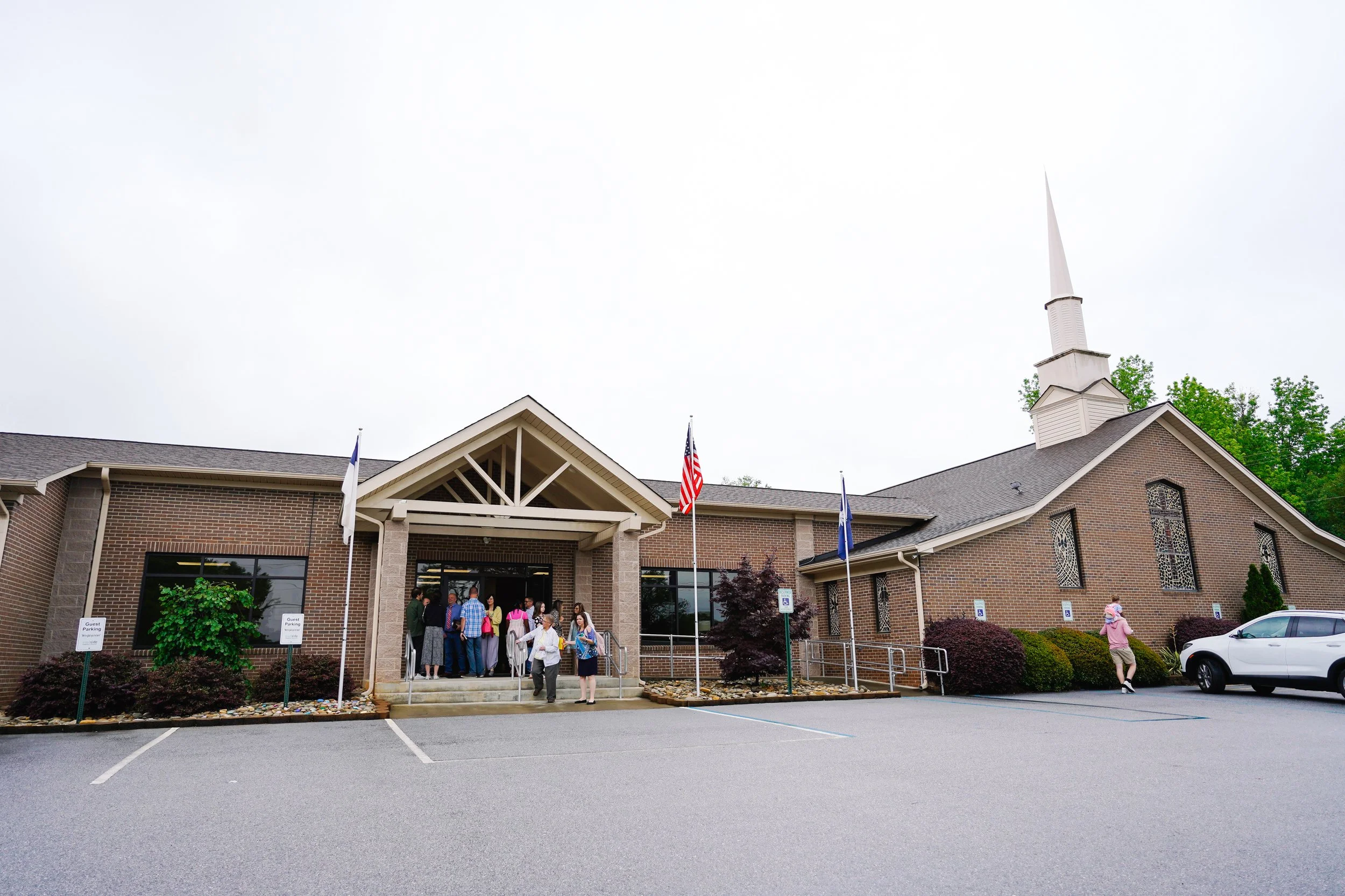 People gathering outside a brick church with a steeple, American flags, and flags of other colors, on a cloudy day.