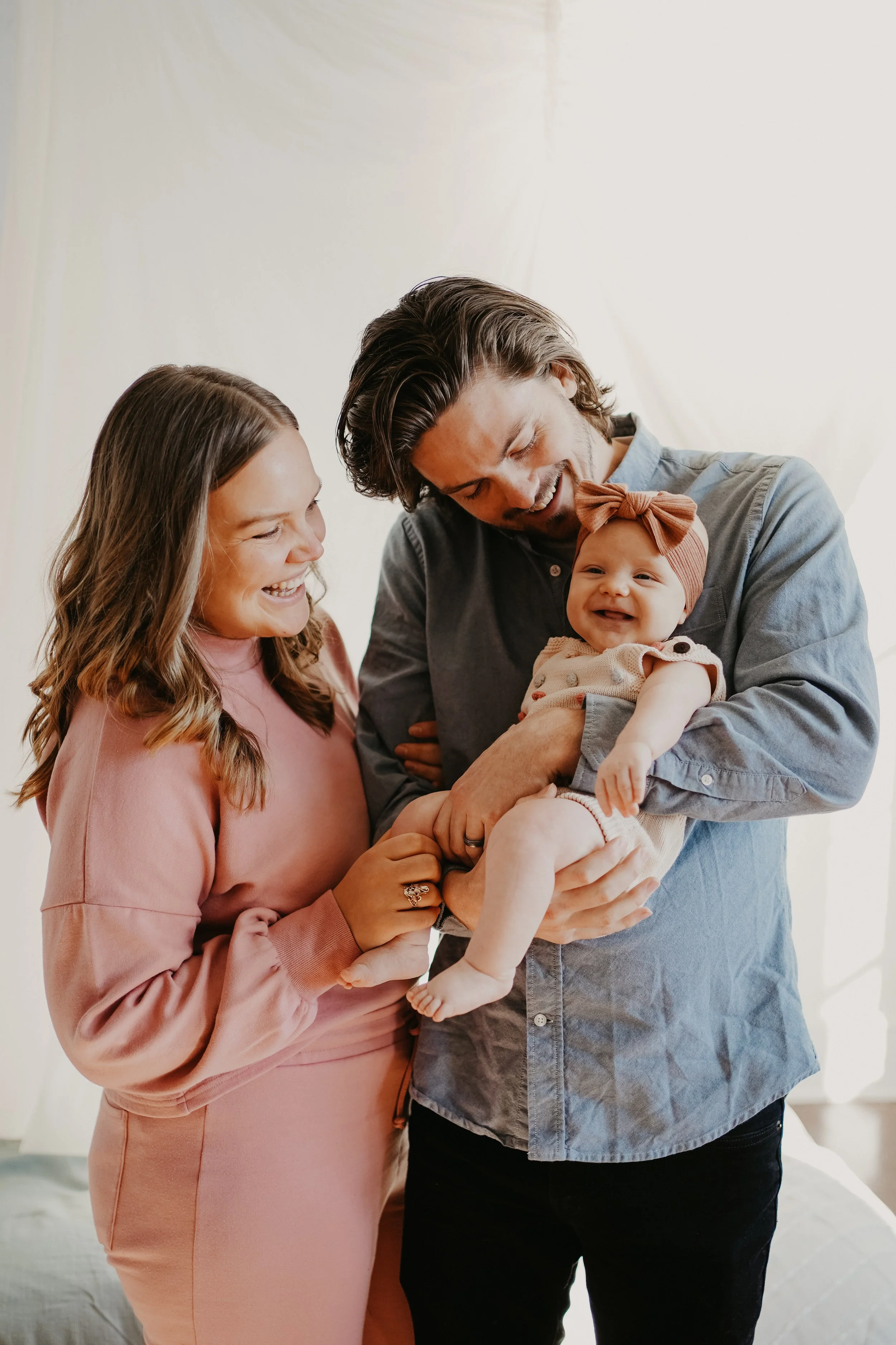 A smiling woman and a smiling man holding a happy baby girl with a bow on her head, in front of a light background.
