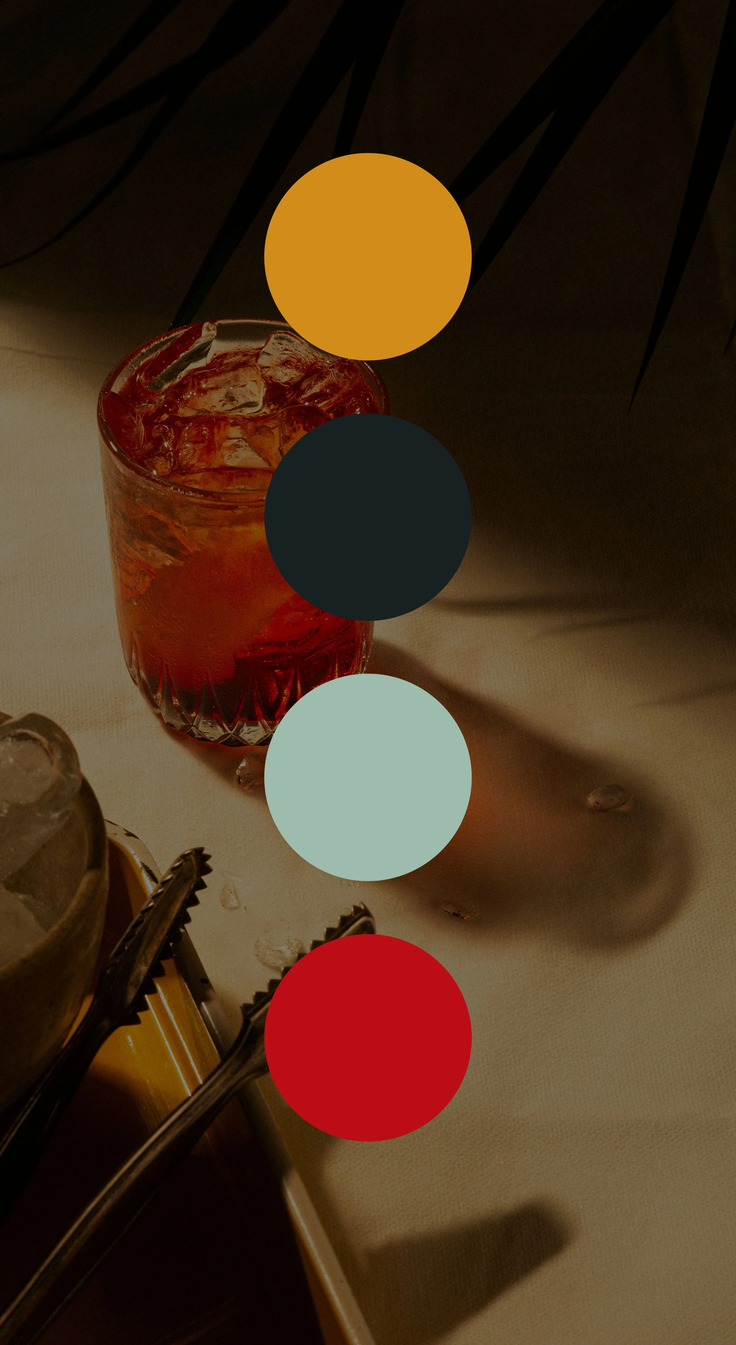 A glass of iced tea with ice cubes on a table, with a shadow cast nearby, and some cocktail tongs resting on a container filled with ice.