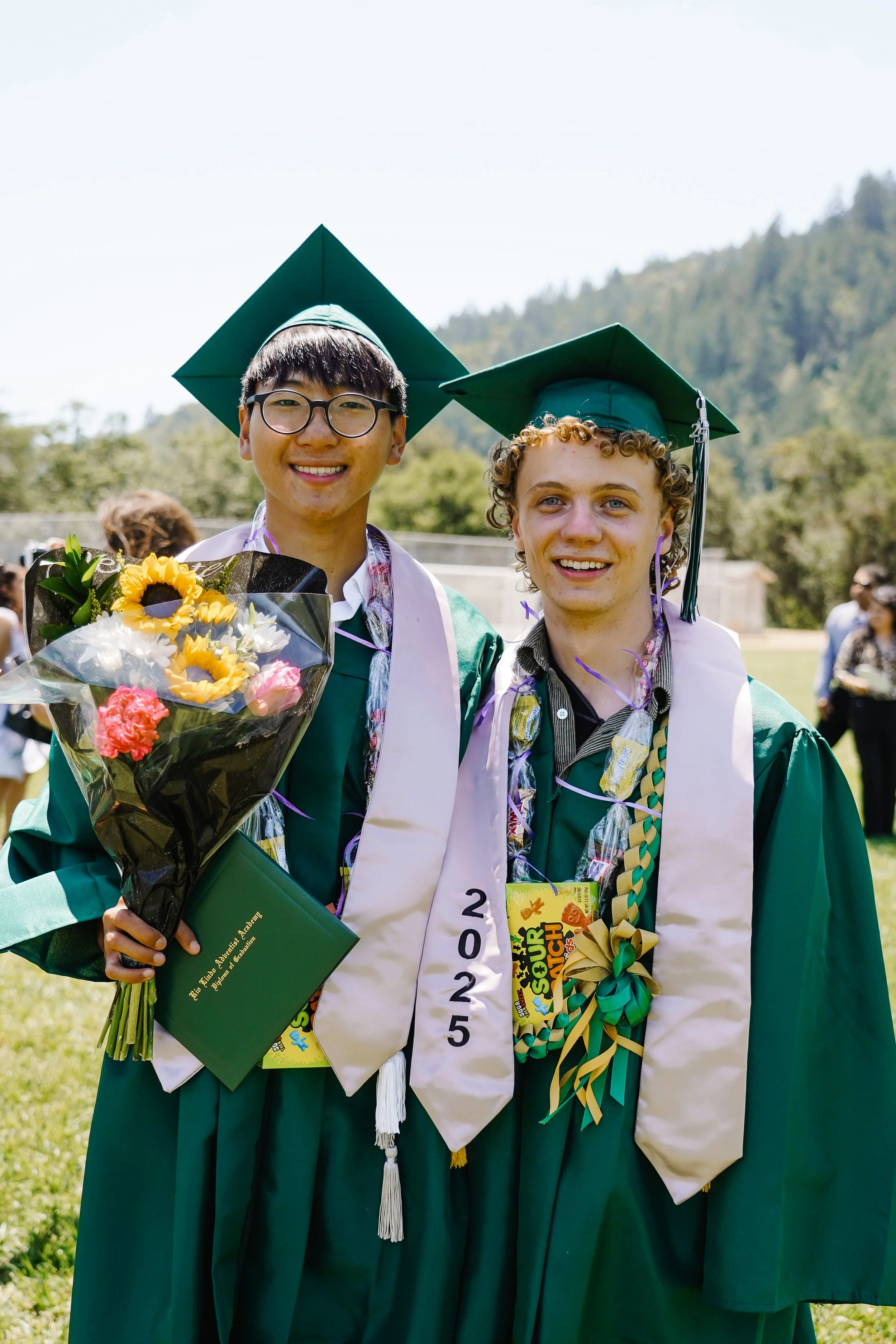 Two graduates in green caps and gowns smiling outdoors, one holding a bouquet of flowers with sunflowers and pink blooms, and both wearing honor cords and sashes, with a mountain and other people in the background.