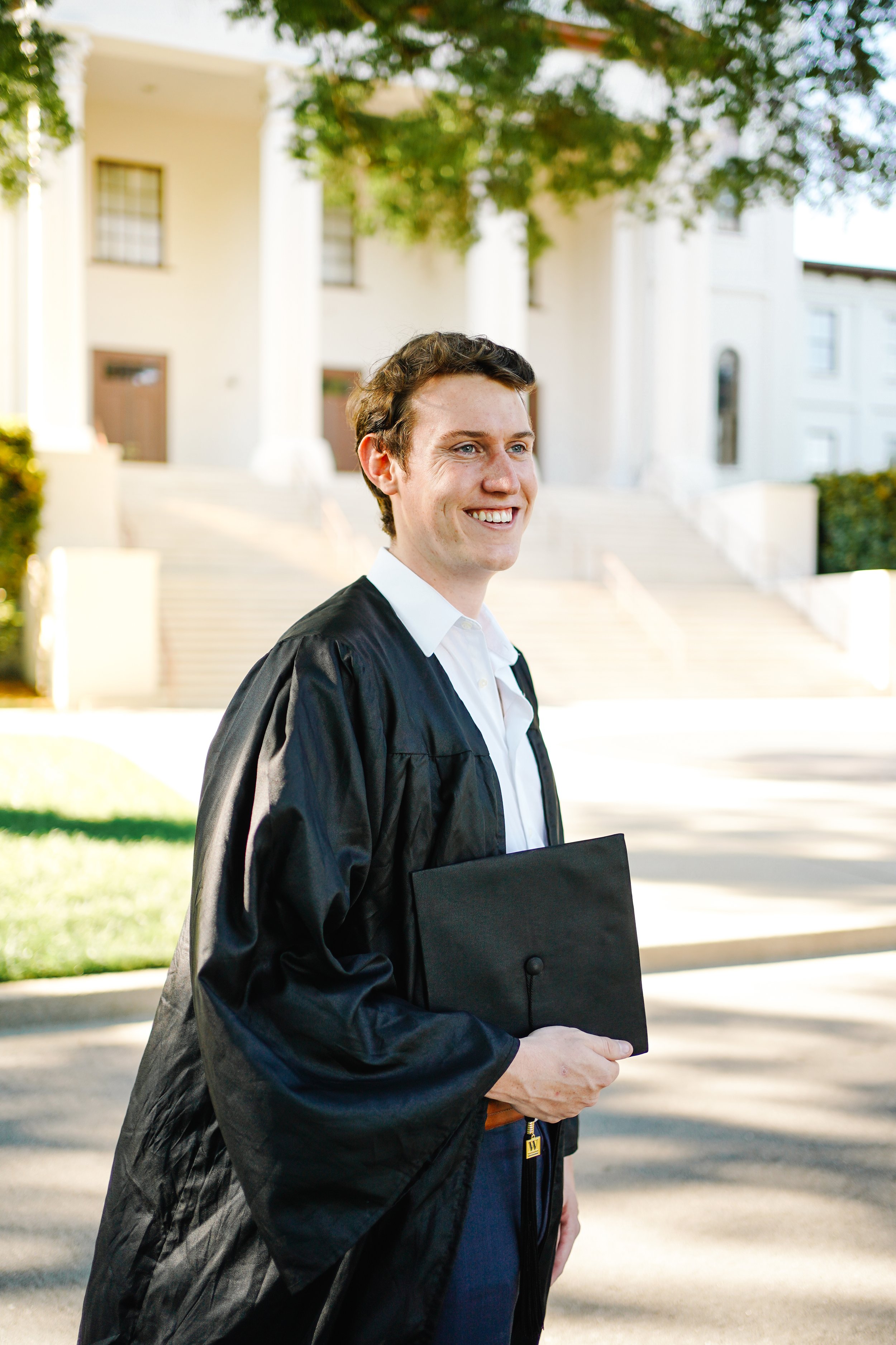 A young man in a graduation gown holding a diploma on the campus with a white building with steps in the background.