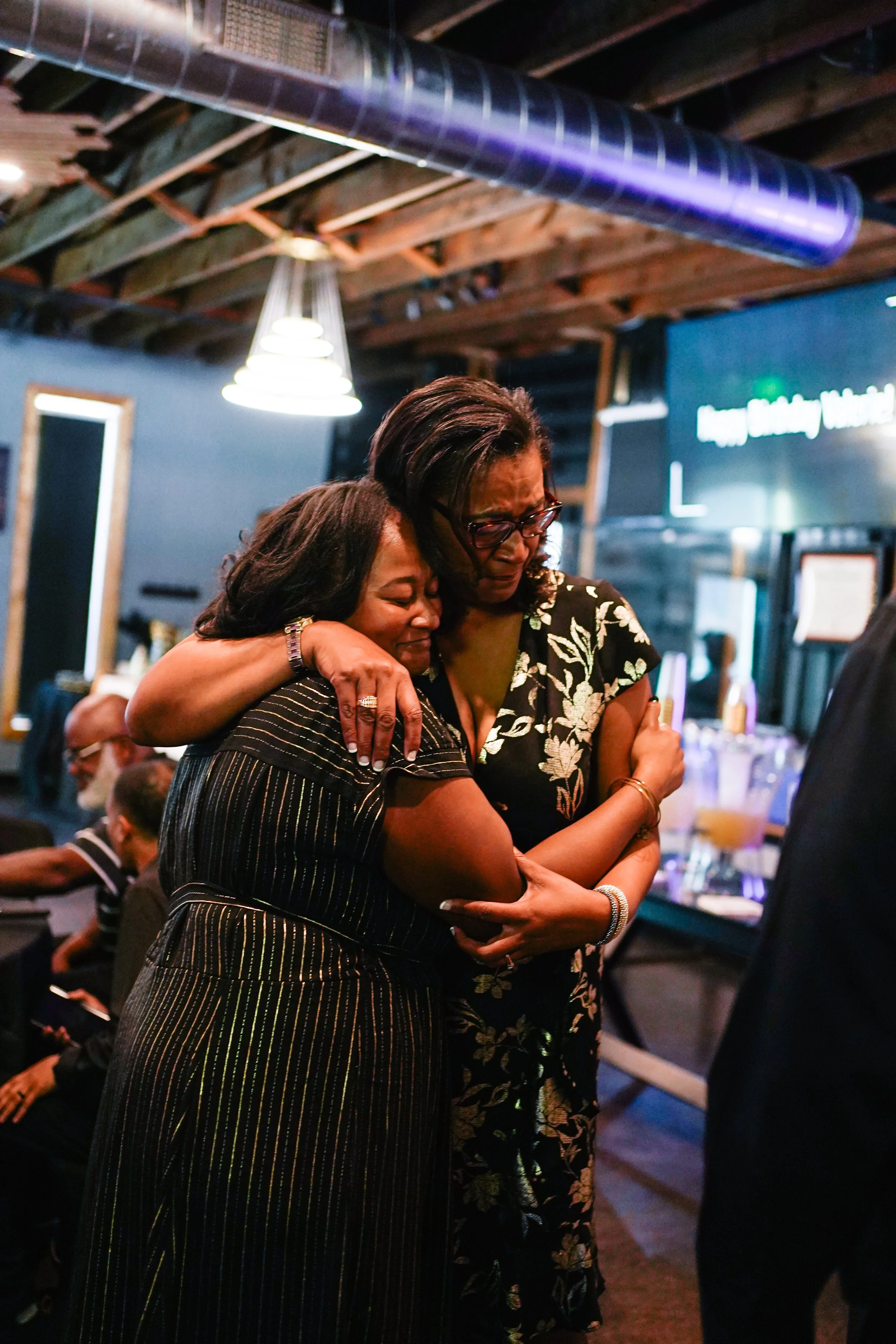 Two women hug each other tightly, displaying emotions of comfort and support, at a social gathering in a dimly lit, rustic indoor setting.