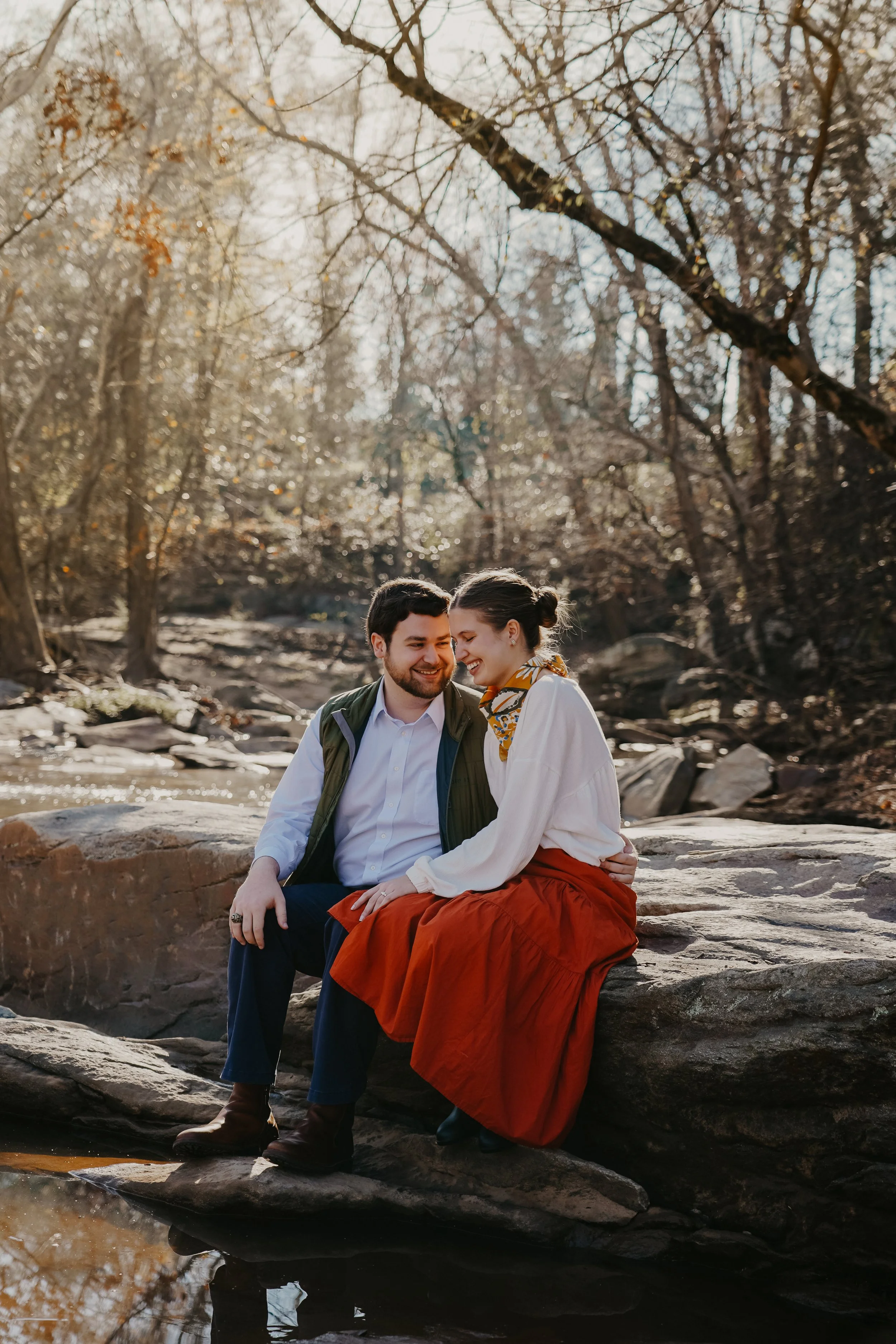 A couple sitting on rocks by a river in a wooded area on a sunny day, smiling and sharing a moment.