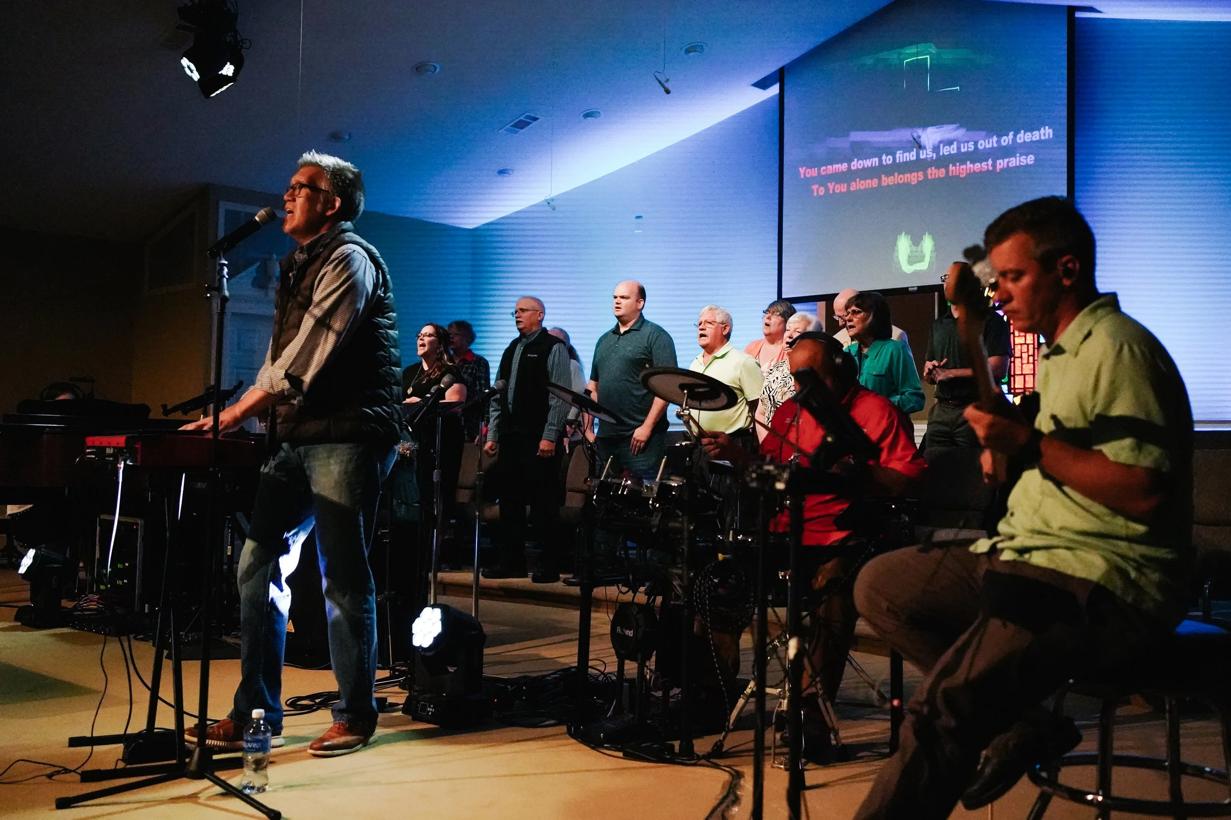 A group of people worshiping and singing on stage, with a musician playing keyboard and others singing and playing instruments, during a religious service with song lyrics displayed on a screen behind them.
