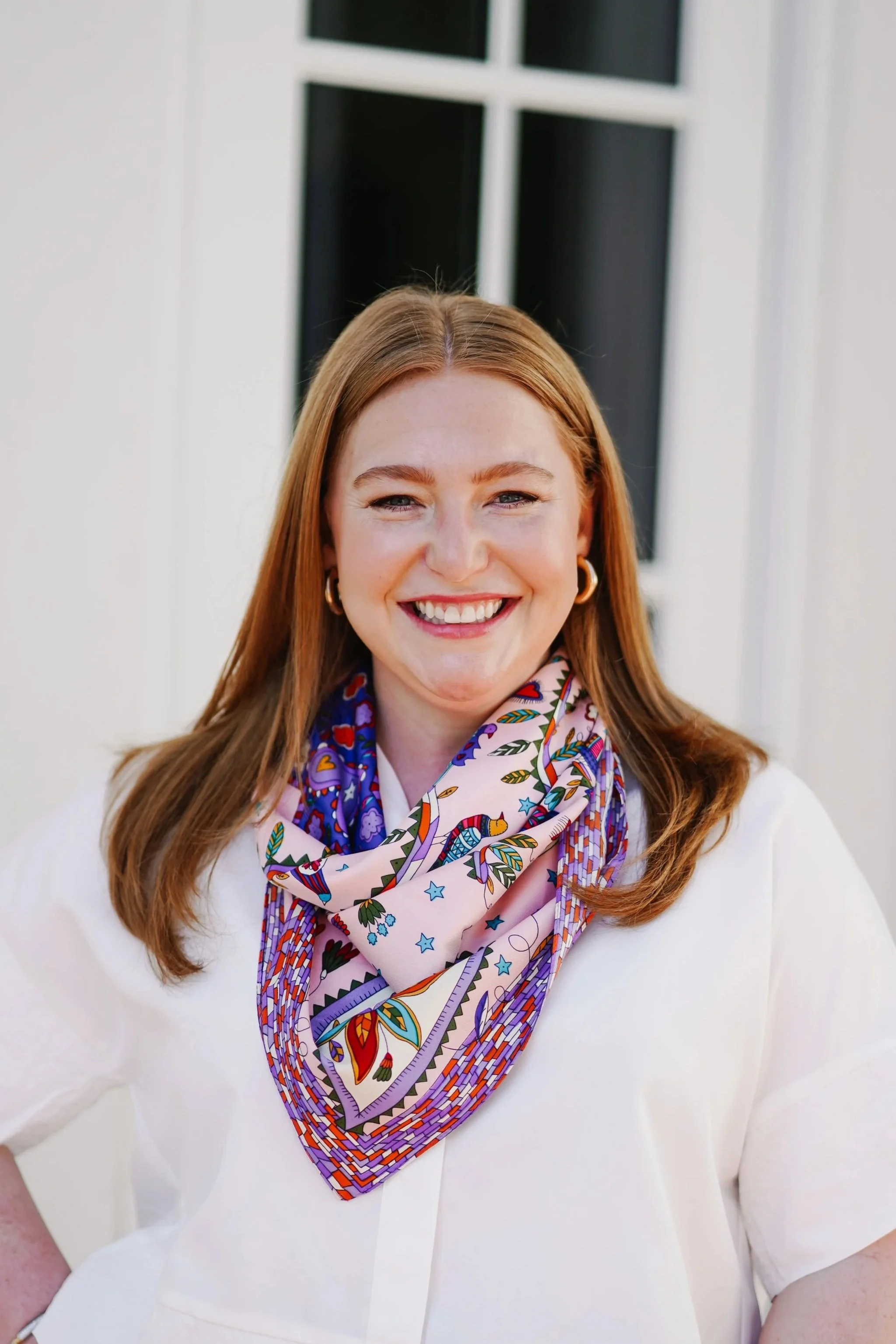 A woman with long red hair, smiling, wearing hoop earrings and a colorful patterned scarf, standing outside in front of a white building with black window panes.