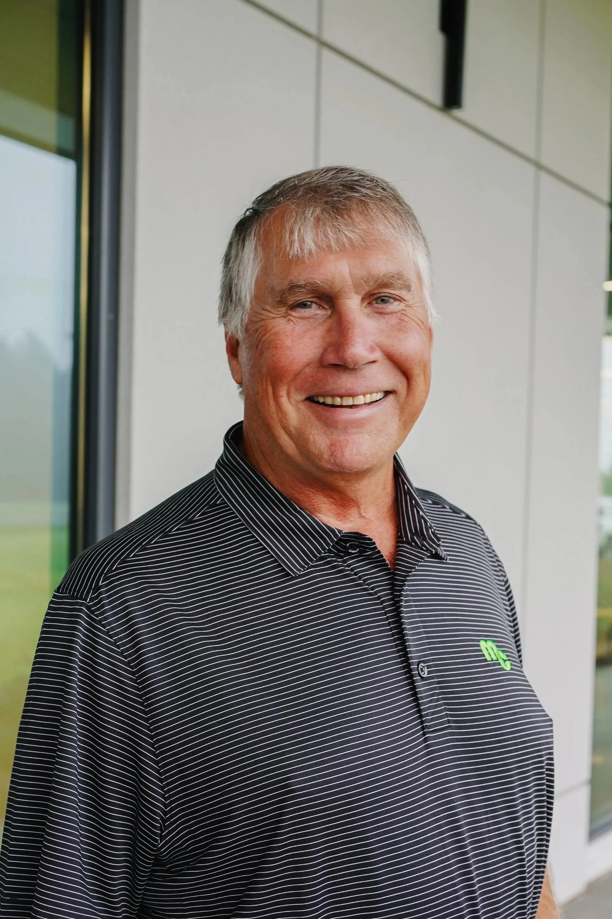 Smiling man in a black and white striped polo shirt standing outdoors near a modern building.