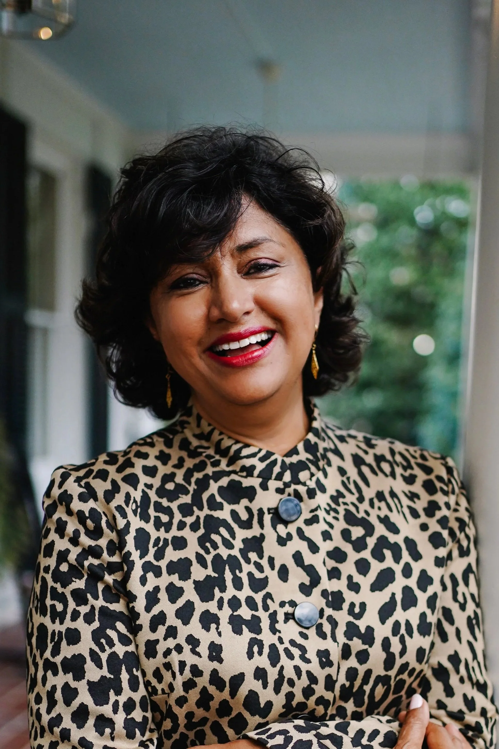 A woman with dark curly hair smiling, wearing a leopard print jacket and gold earrings, standing outdoors.