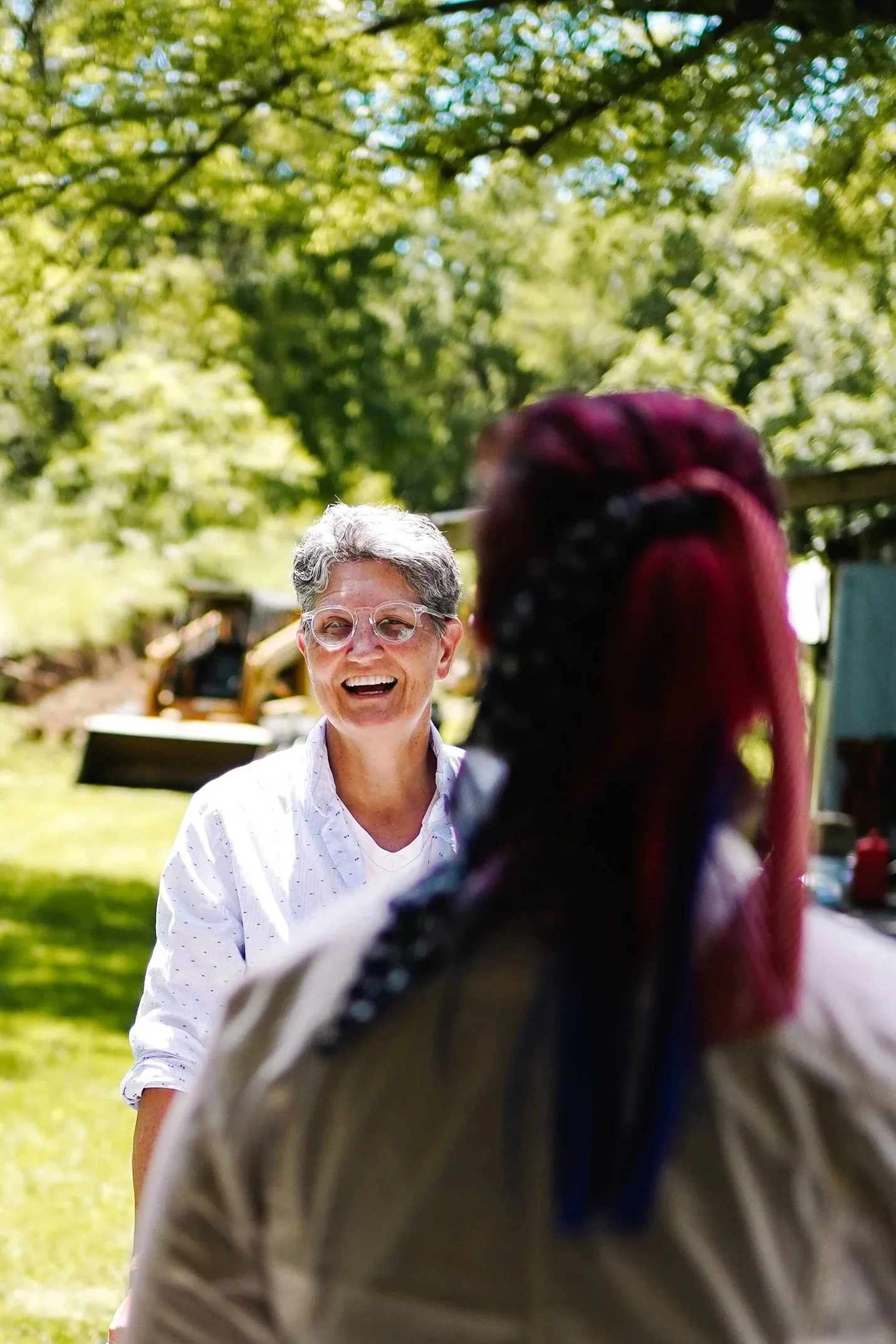 Two women having a conversation outdoors in a park with green trees; one woman with short gray hair, glasses, and a white shirt laughing, the other woman with long multicolored braids, seen from behind.