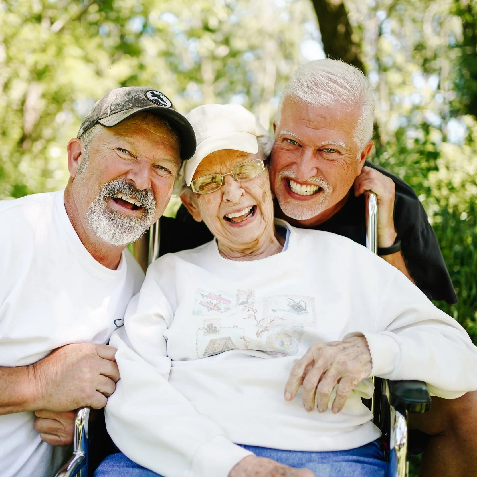 Three older adults smiling happily outdoors in a green park, with one woman in a wheelchair and two men leaning in close.