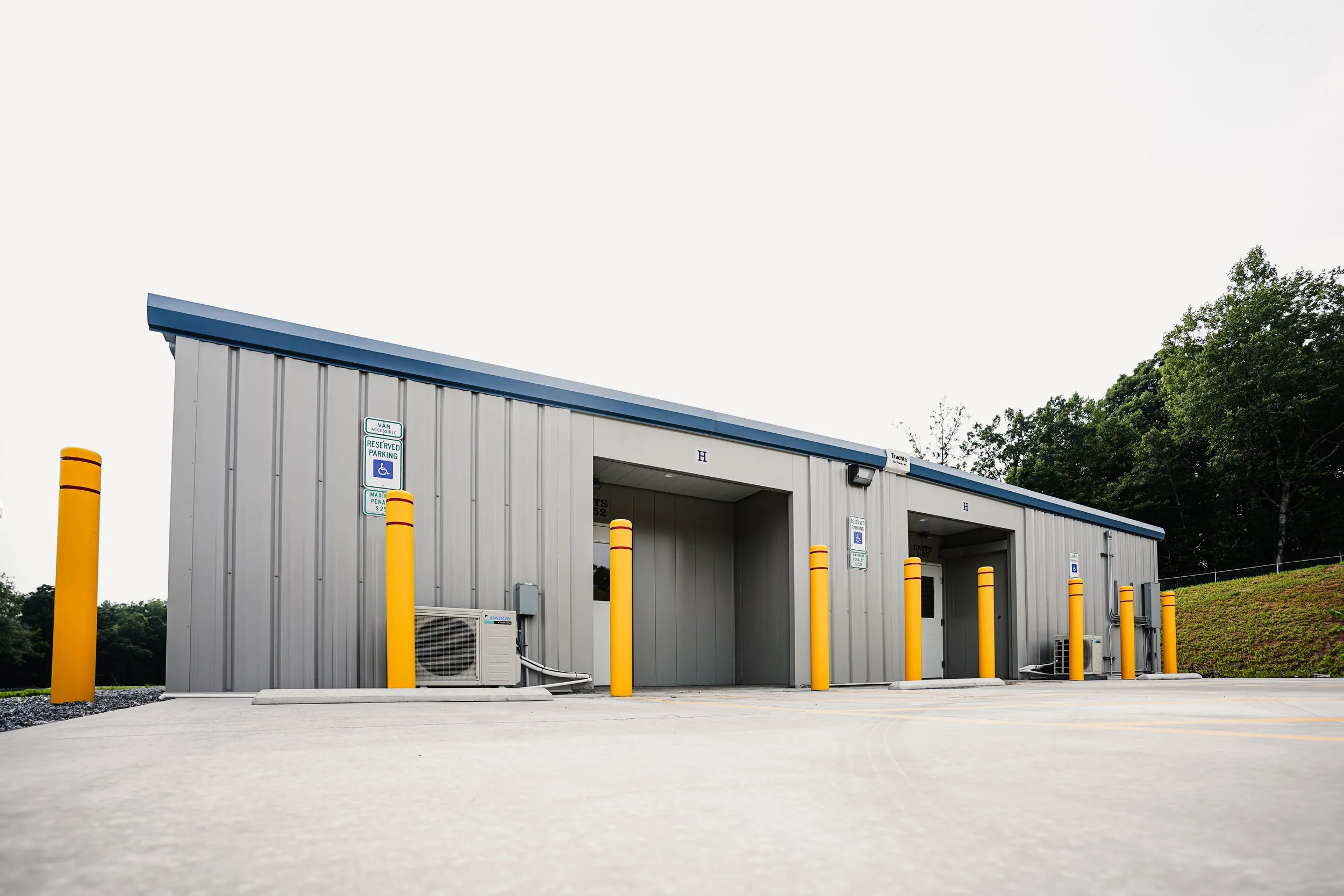 A small, gray metal building with multiple parking spaces in front, marked for handicapped parking and reserved parking. Yellow safety bollards surround the parking spaces, and there are two air conditioning units on the exterior wall. The building is located on a paved area with a grassy hill and trees in the background.
