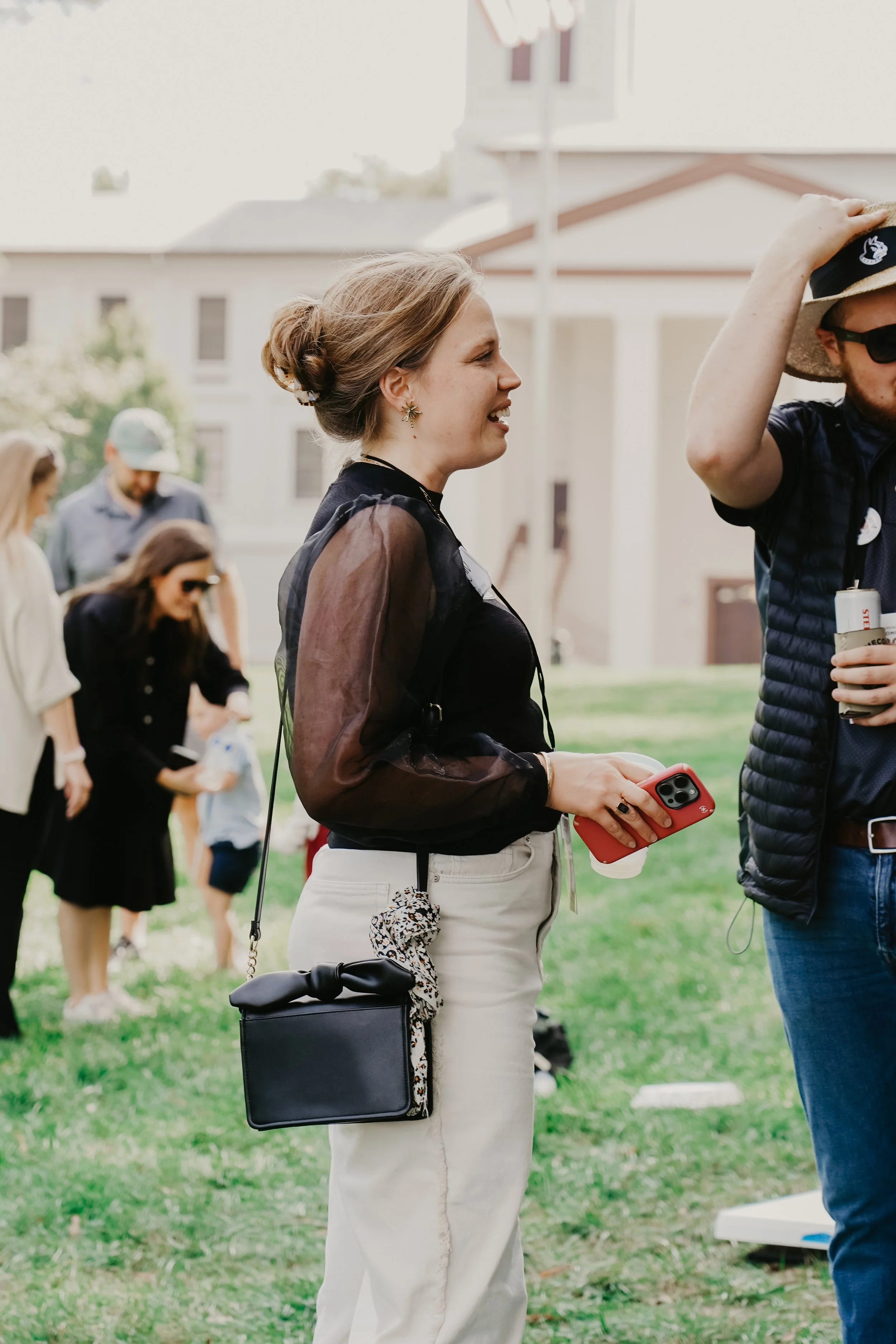A woman with blonde hair tied in a bun stands outdoors at a social gathering, holding a smartphone in her right hand. She wears a black sheer blouse and white pants, accessorized with earrings, a black handbag with a patterned scarf tied to its handl