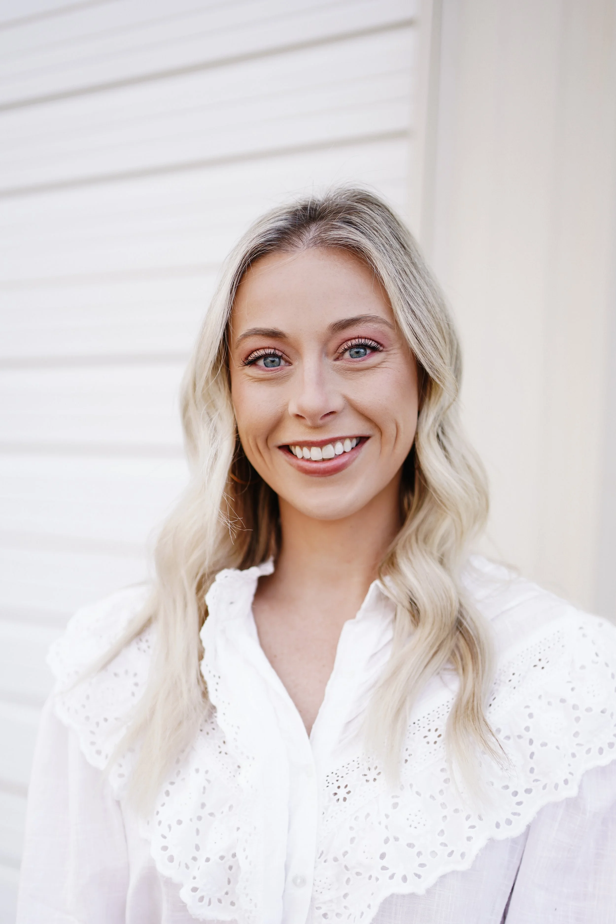 A young woman with blonde wavy hair, blue eyes, and a white eyelet blouse, smiling outdoors against a light-colored, horizontal siding wall.