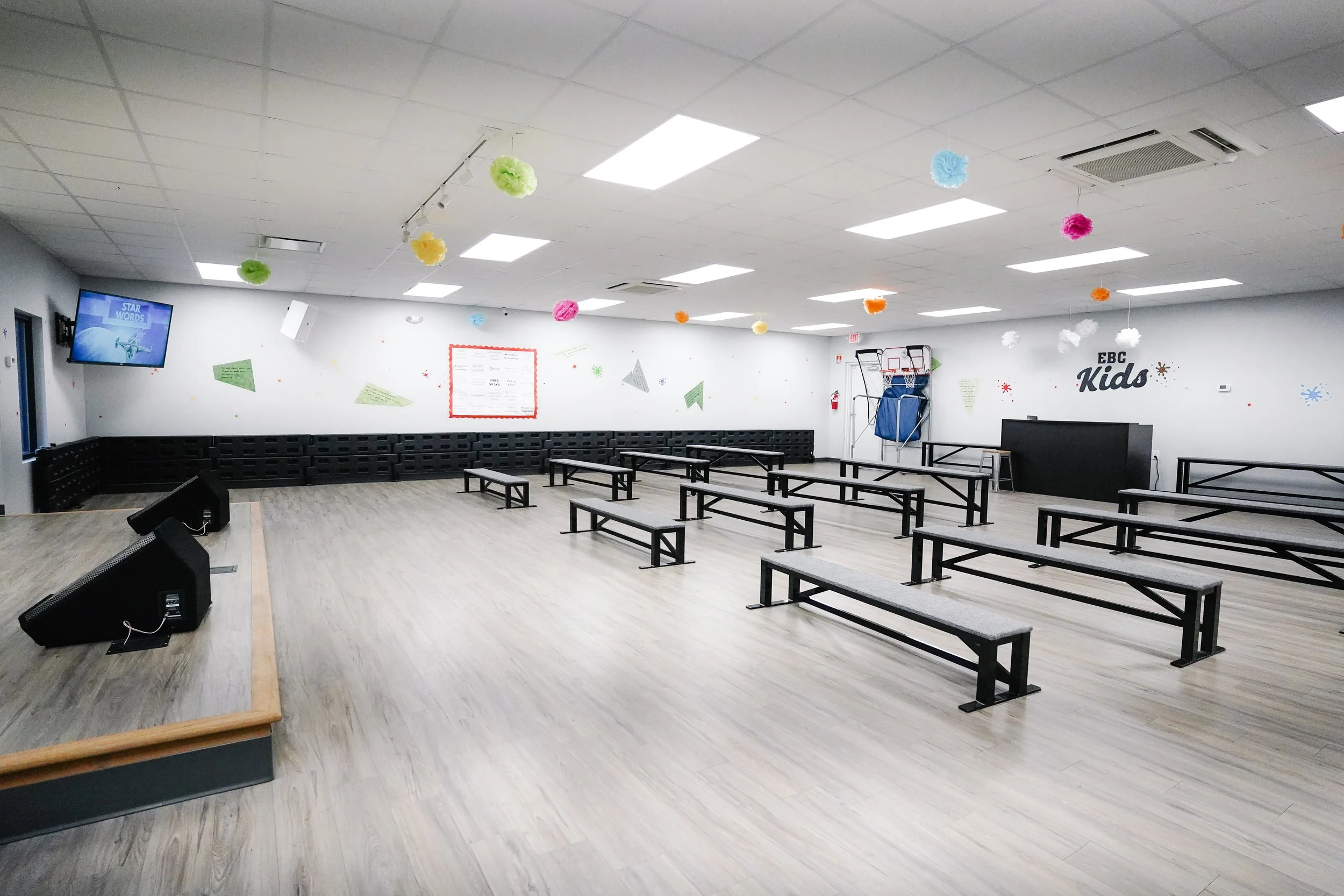 Empty indoor space with rows of benches, a small stage with speakers, and colorful paper decorations hanging from the ceiling, labeled as EBC Kids.