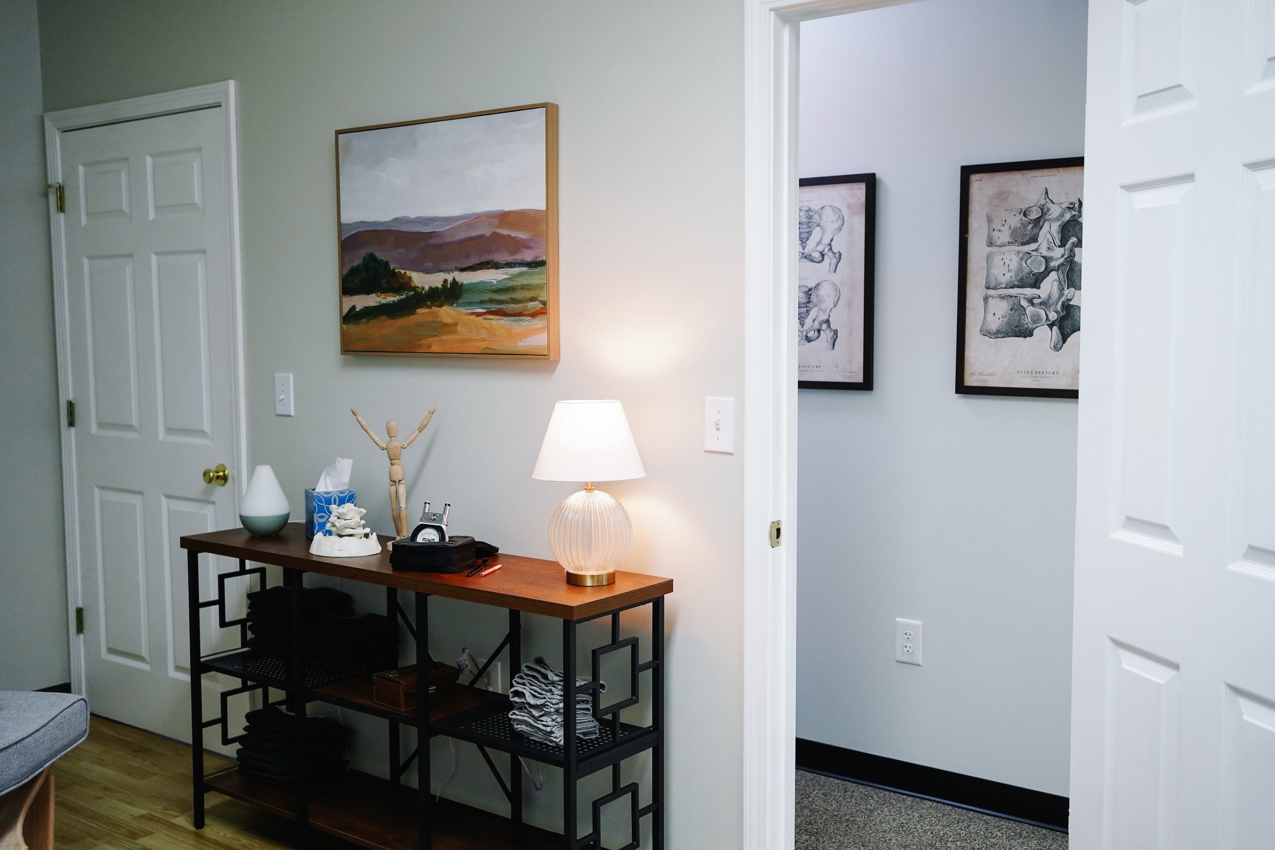 Interior view of a home with a wooden console table decorated with a lamp, tissue box, a decorative hand, and a clock. Above the table is a landscape painting. A hallway with frameless sketches of animal bones is visible through an open door.