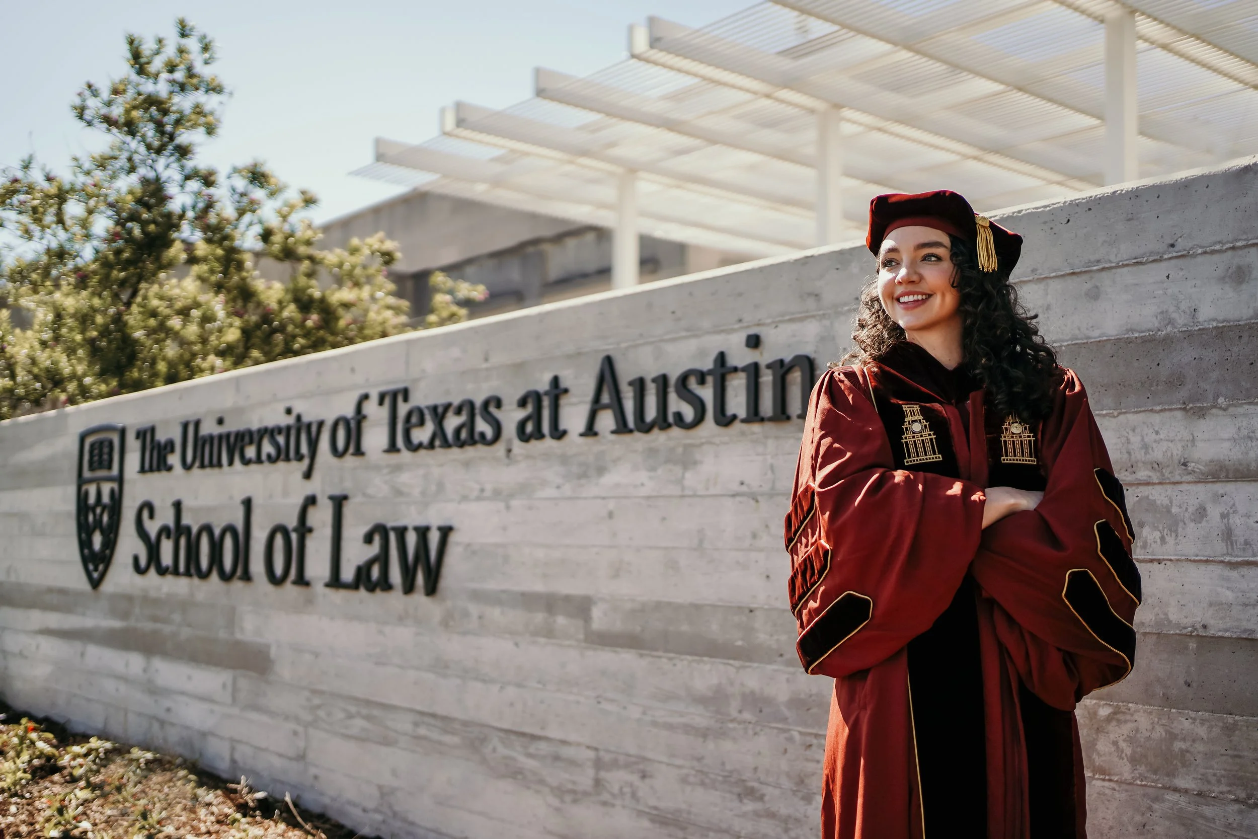 Graduate woman wearing maroon and black cap and gown standing outside in front of a sign for the University of Texas at Austin School of Law, smiling with arms crossed.