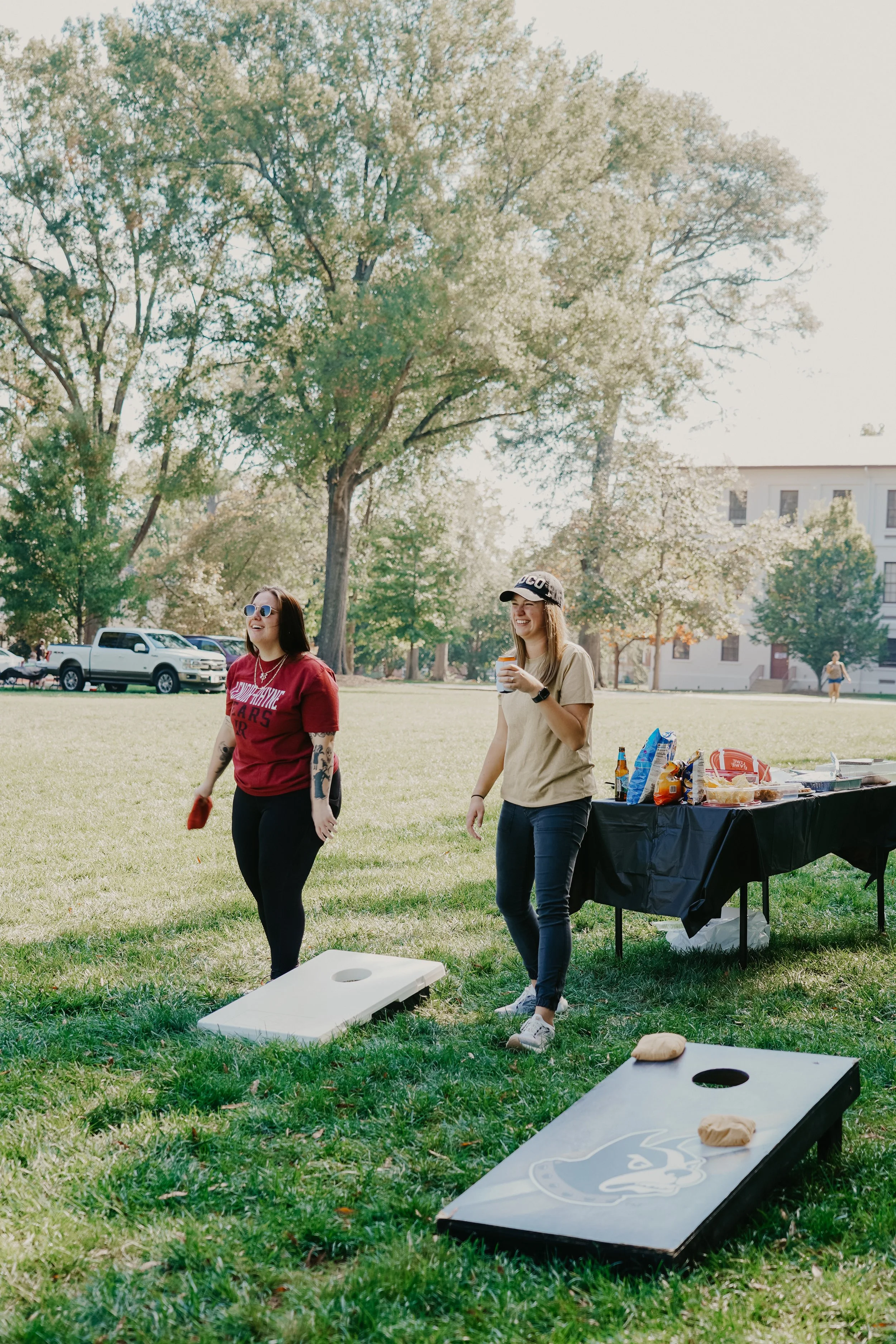 Two women playing cornhole at a park during daytime, with a table of snacks and drinks behind them and a large tree and building in the background.