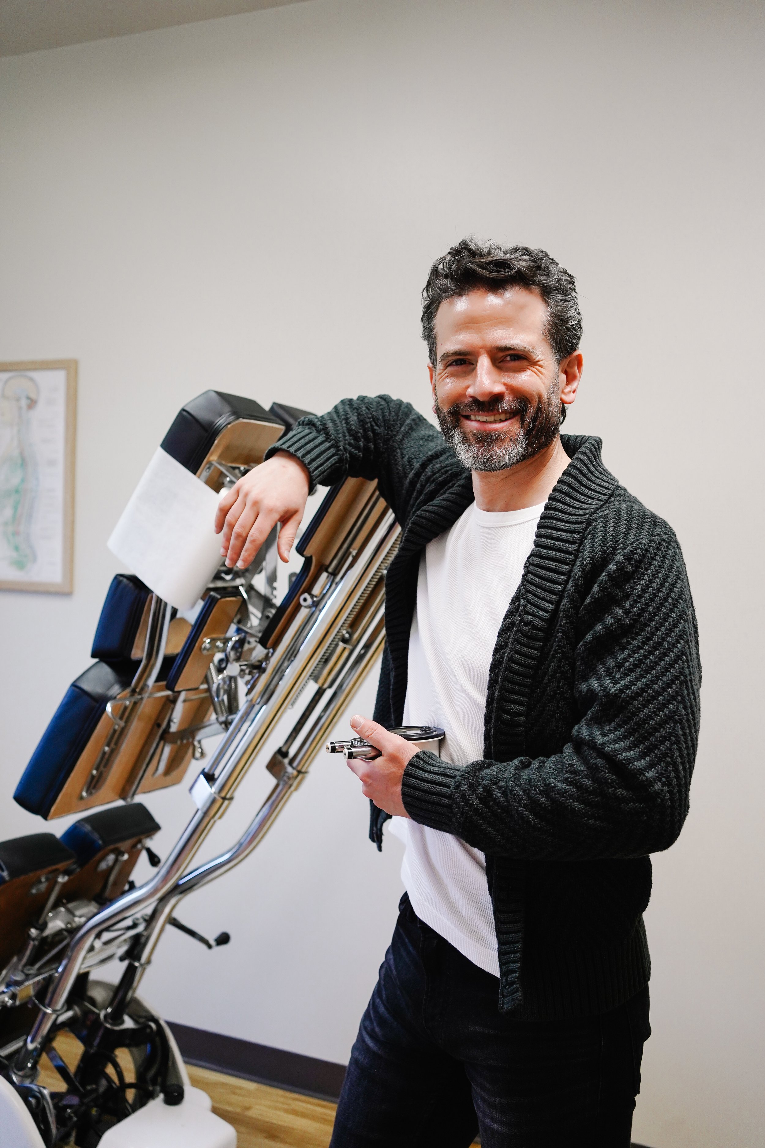 A smiling man with dark hair and a beard leaning on a medical or dental examination chair in a clinic or office setting.