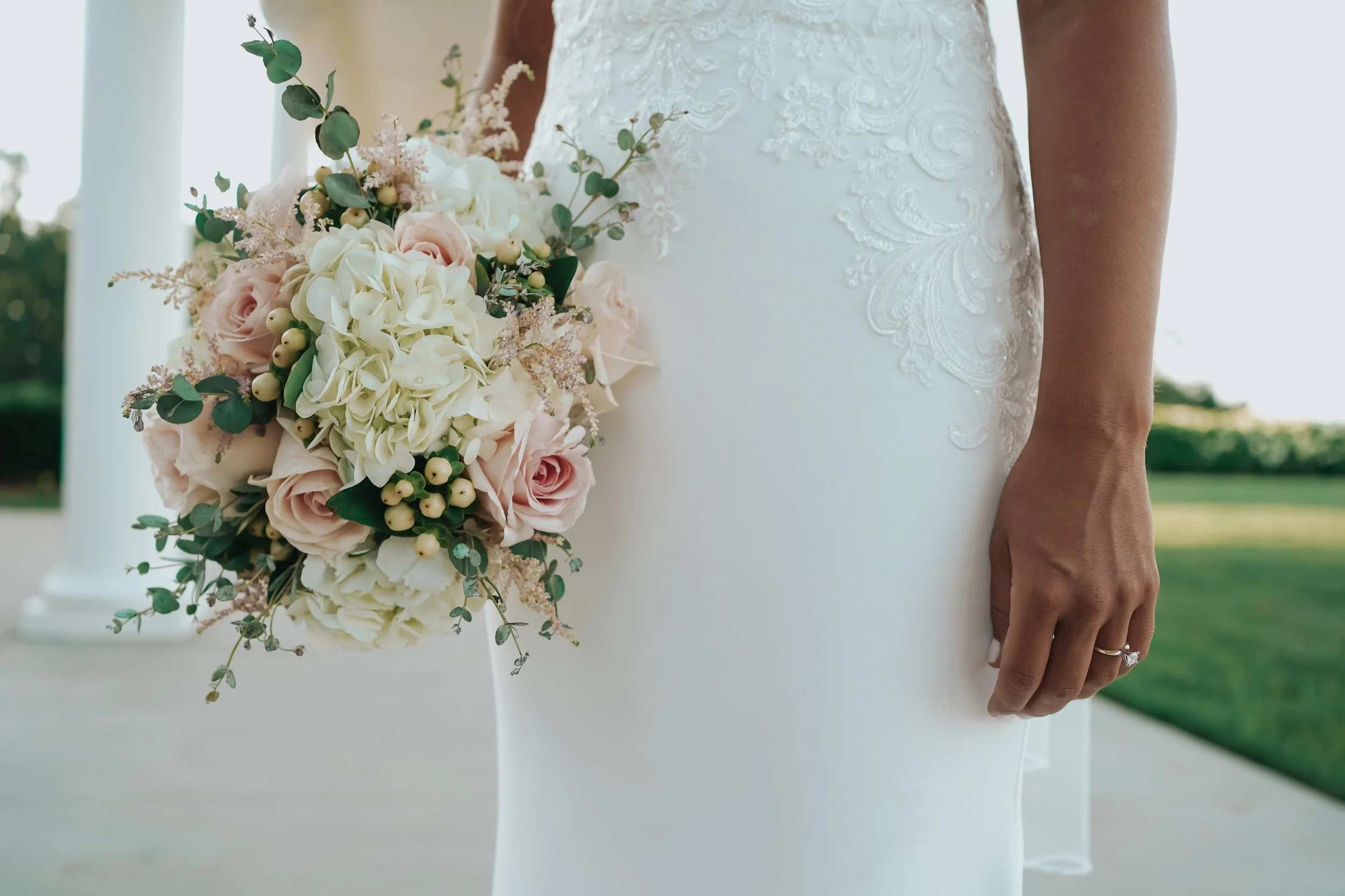 Close-up of a bride holding a bouquet of pink roses, white hydrangeas, and greenery, wearing a white wedding gown with lace details.